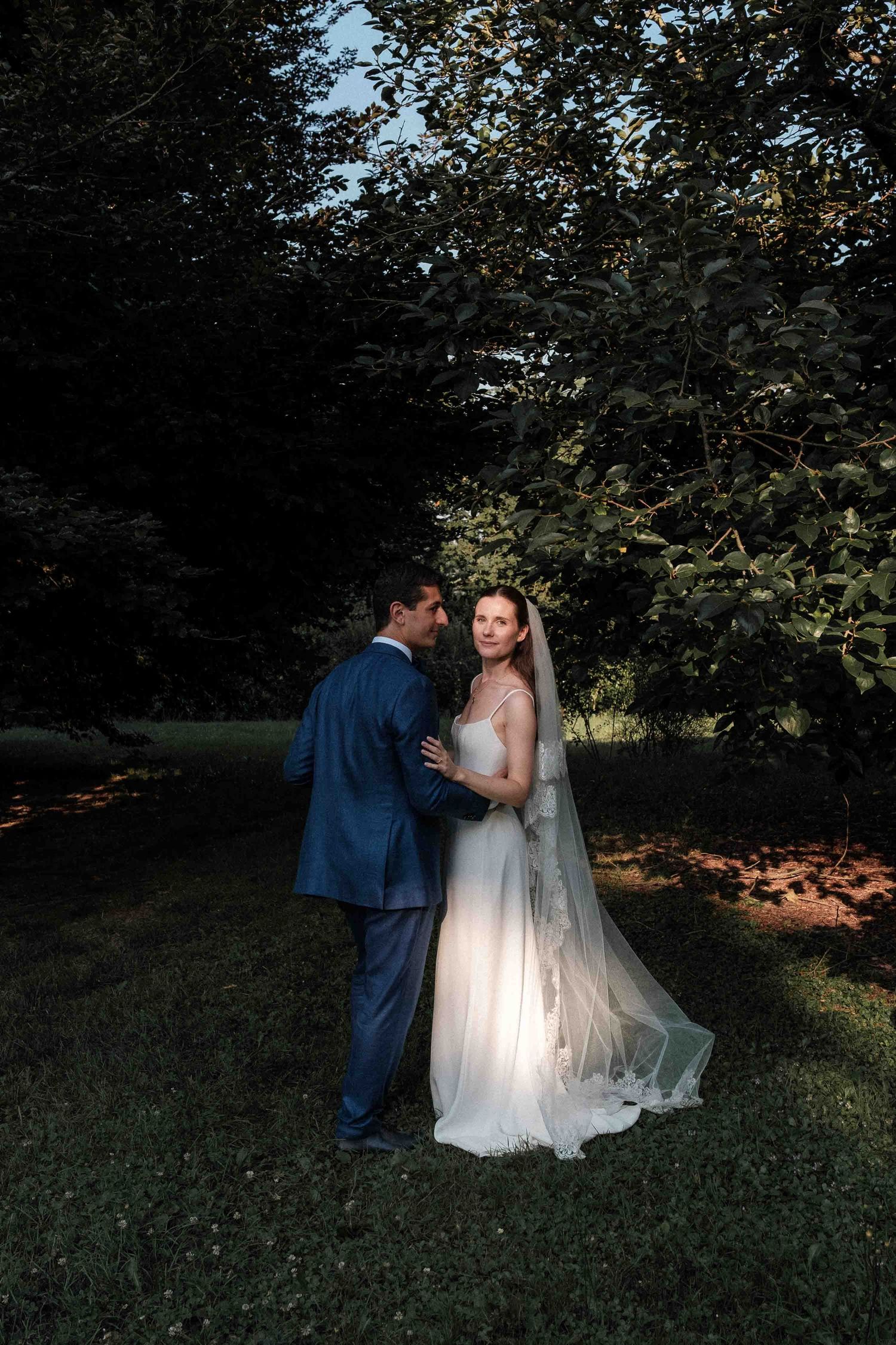 A romantic wedding couple embraces in a sunlit garden setting with trees in the background.