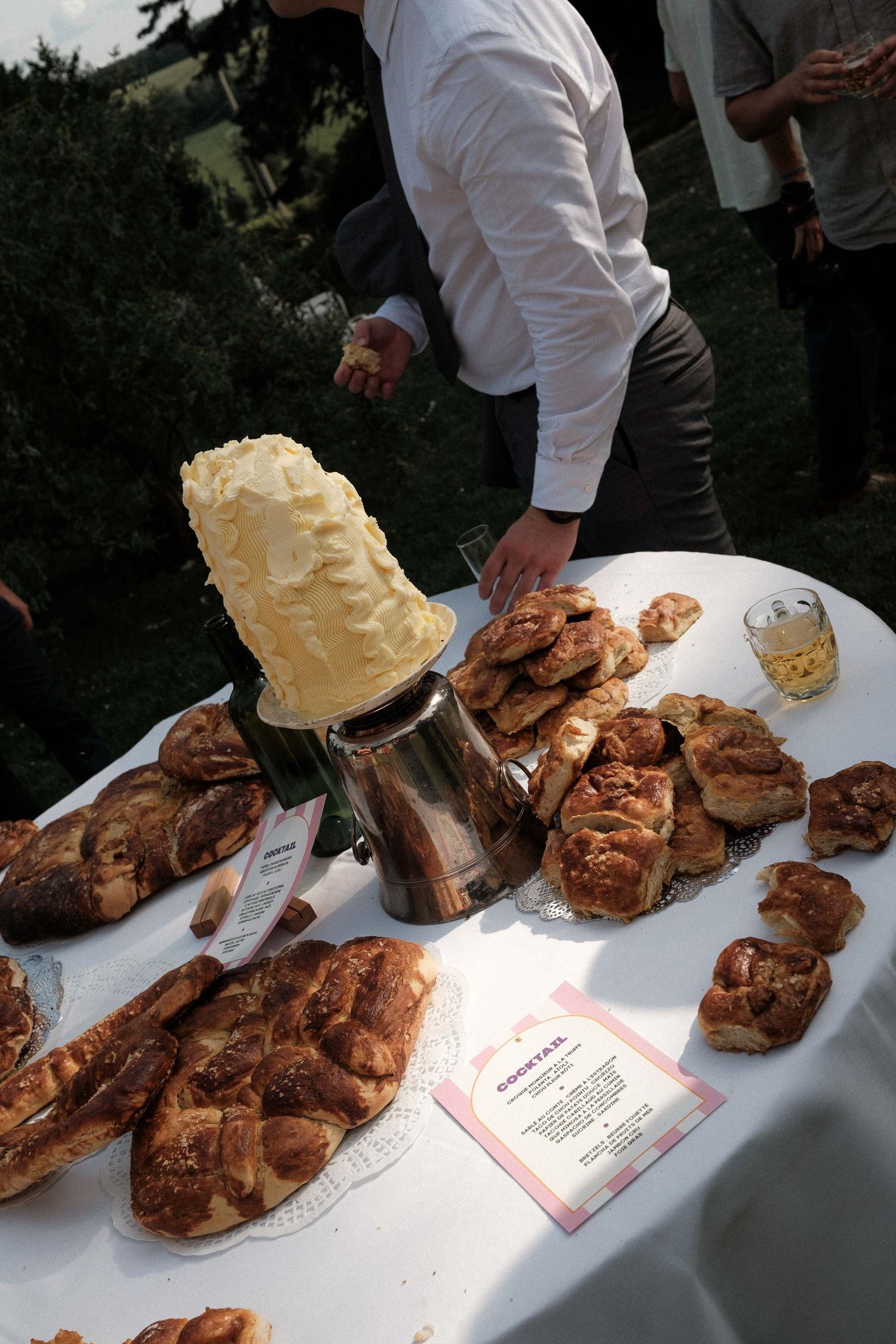 Catered event setup showing platters of grilled chicken and whipped butter on a white tablecloth outdoors.