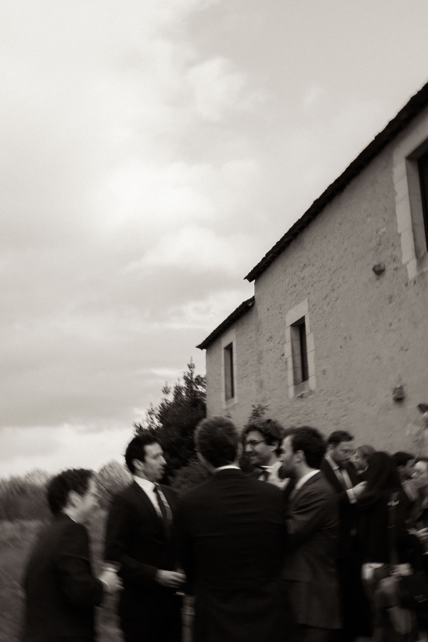 A black and white photograph shows a group of people gathered outside a white building under an overcast sky.