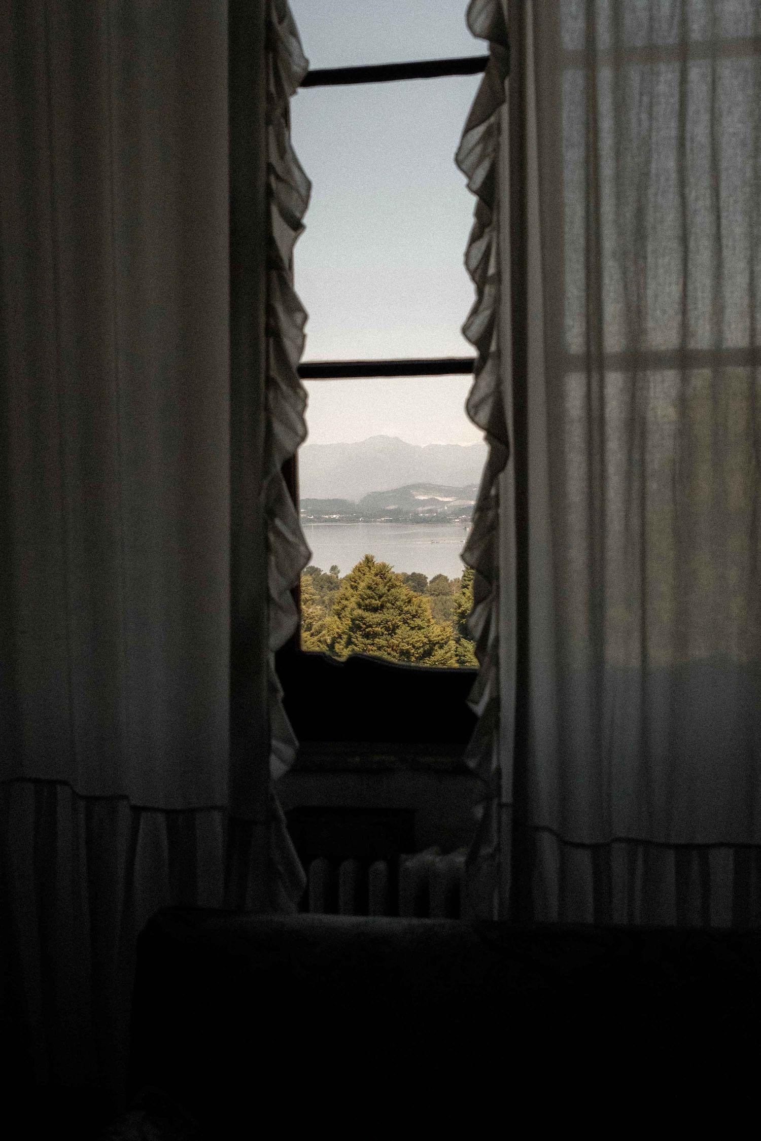 View through parted curtains of a scenic mountain landscape and water visible from a window at dusk.