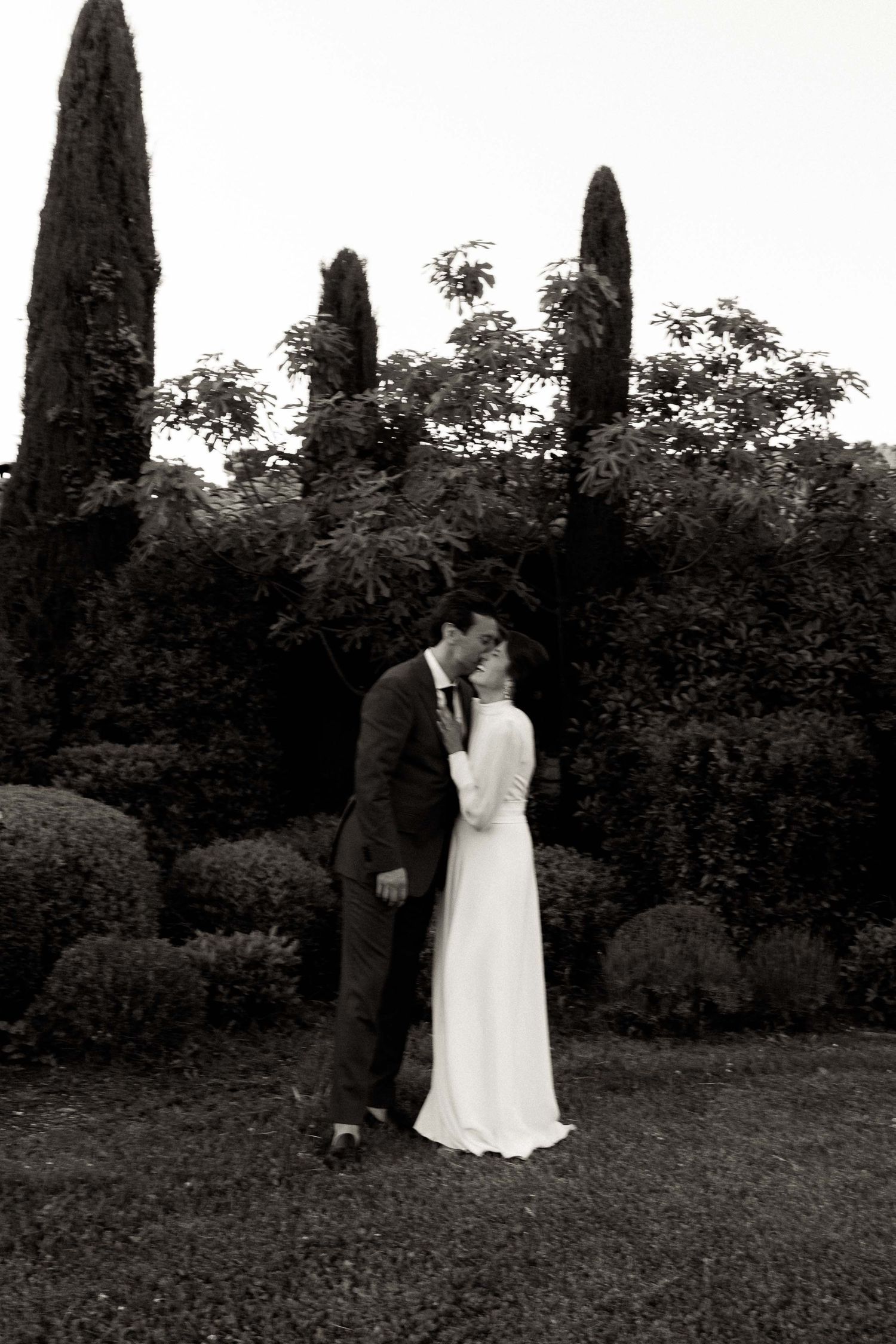 A couple shares an intimate moment in a garden setting surrounded by towering cypress trees.
