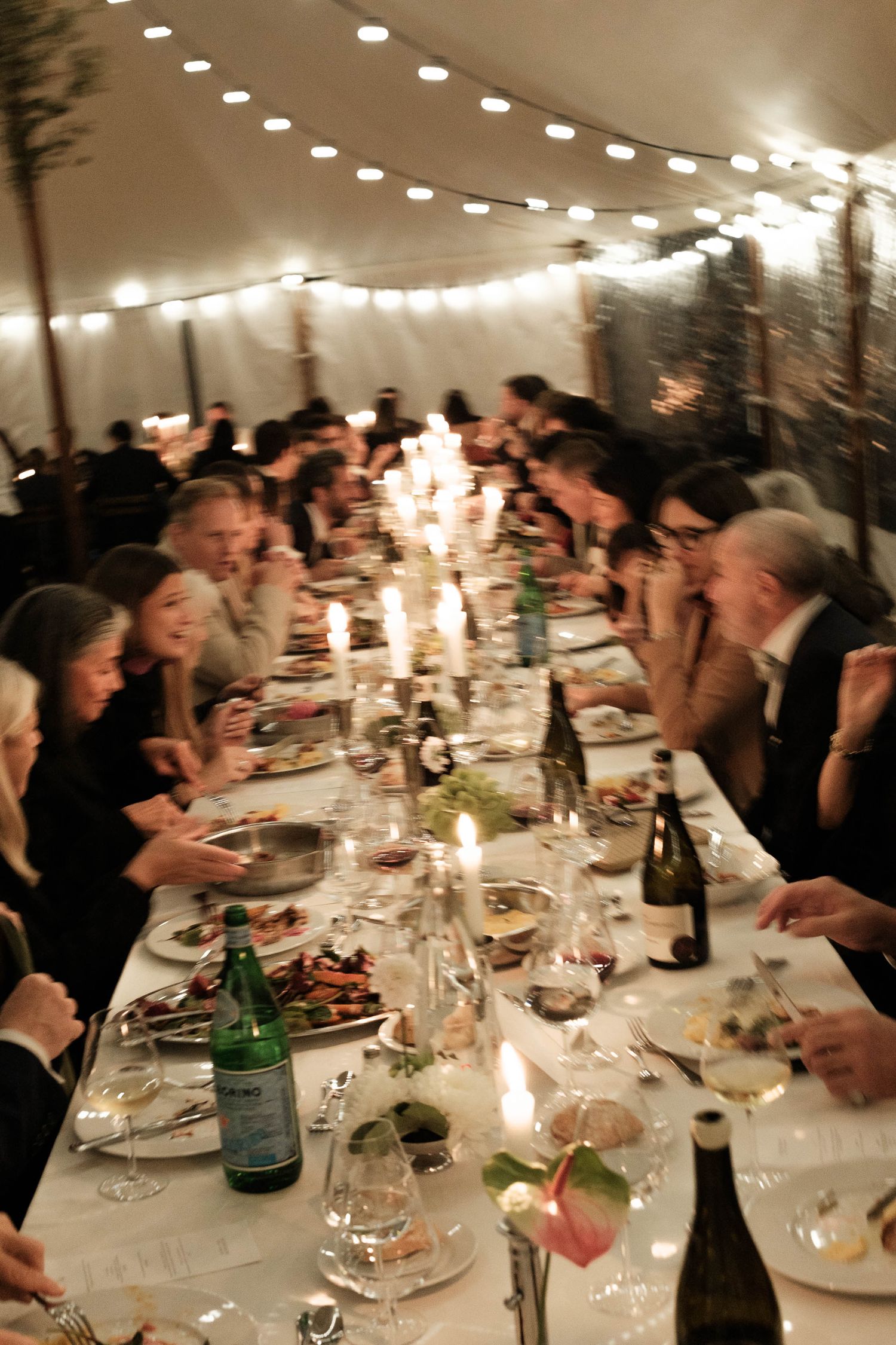 A long dining table filled with guests at an elegant evening reception under warm lighting and string lights.