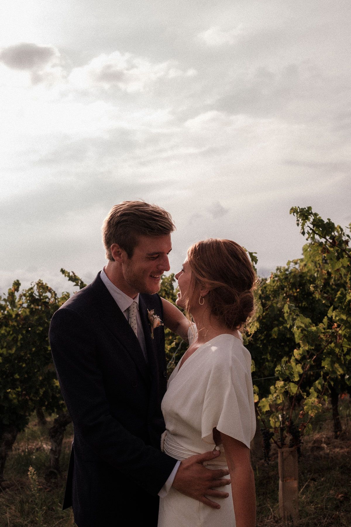 A romantic couple shares an intimate moment in a vineyard at sunset with moody clouds in the background.
