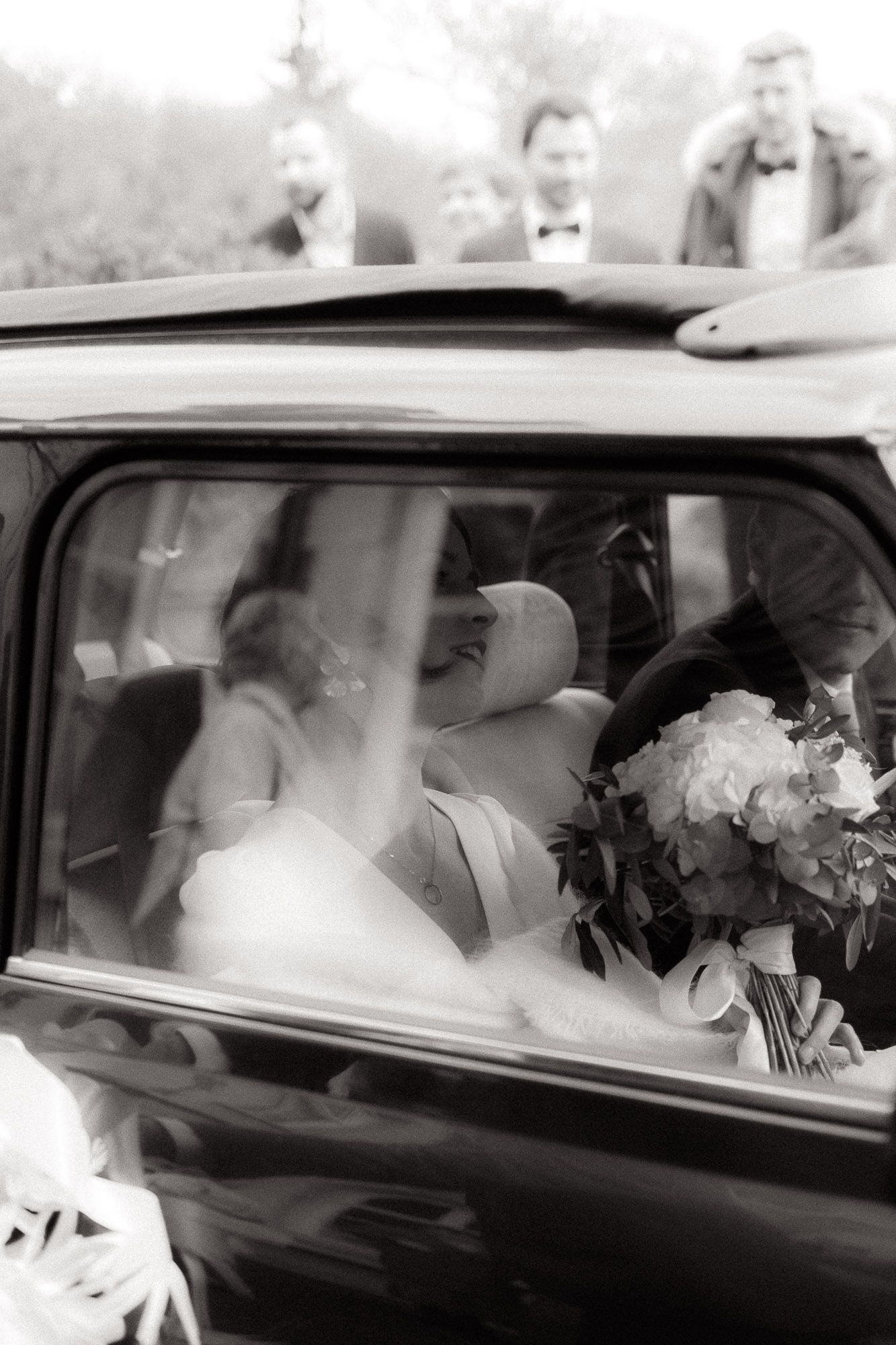 A bride in a white wedding dress sits in a classic vintage car holding a floral bouquet.