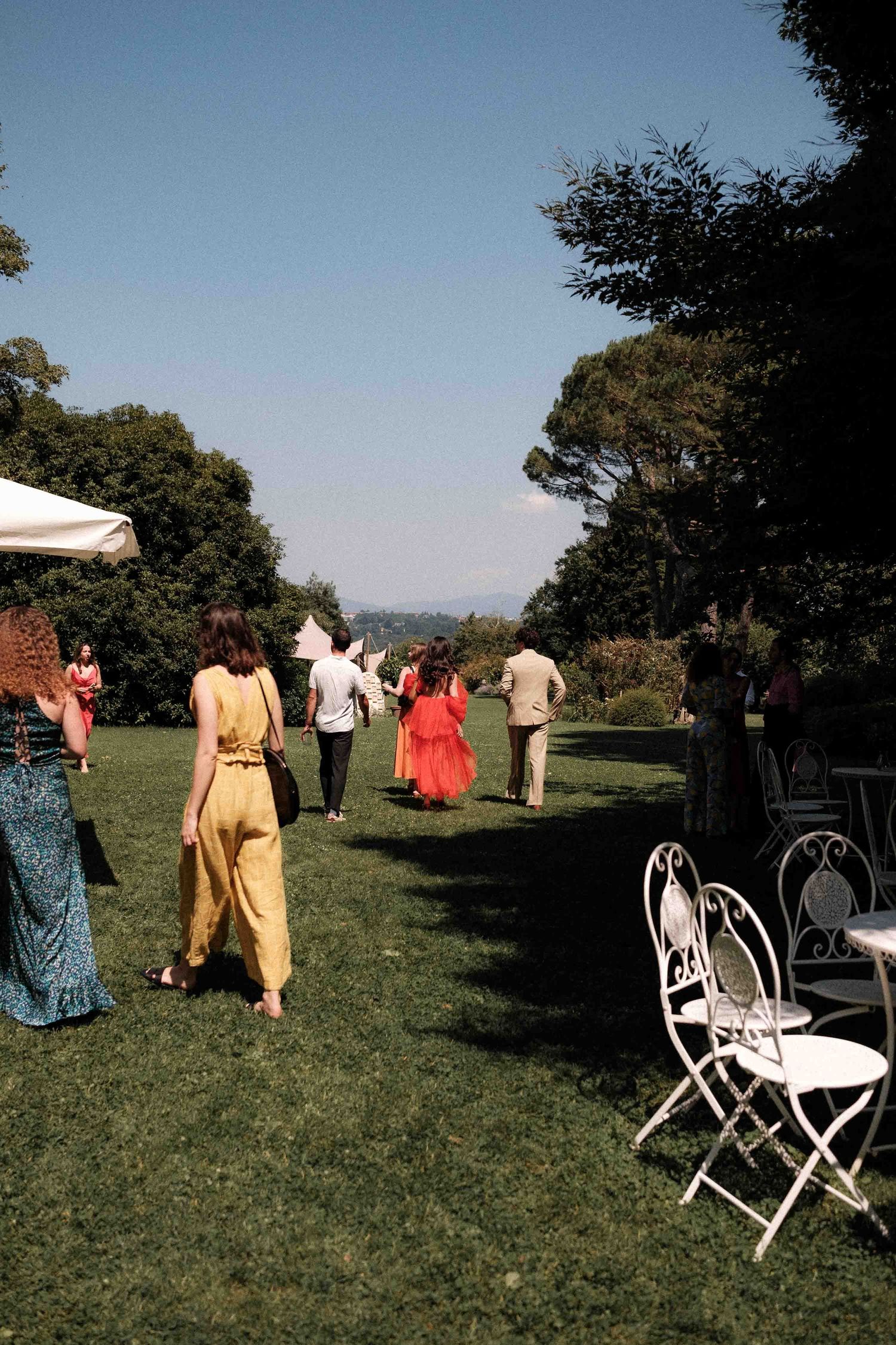 Wedding guests in colorful attire walking across a sunny lawn with white chairs set up outdoors.
