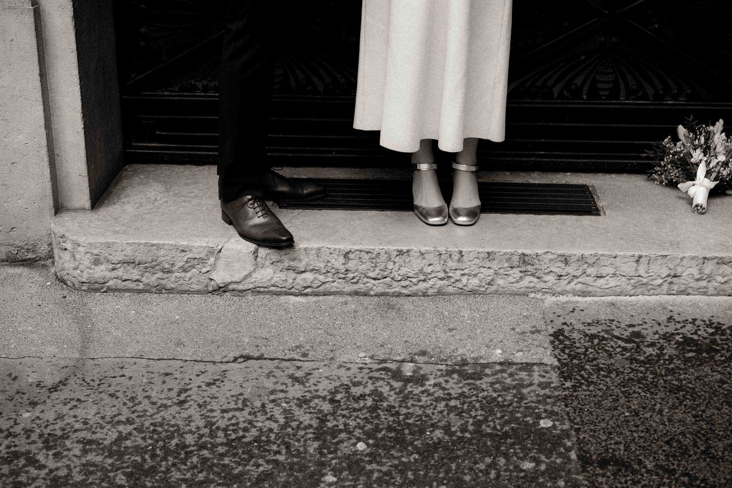 Black and white photograph showing two pairs of feet at a doorway threshold, one in dress shoes and one in white shoes.
