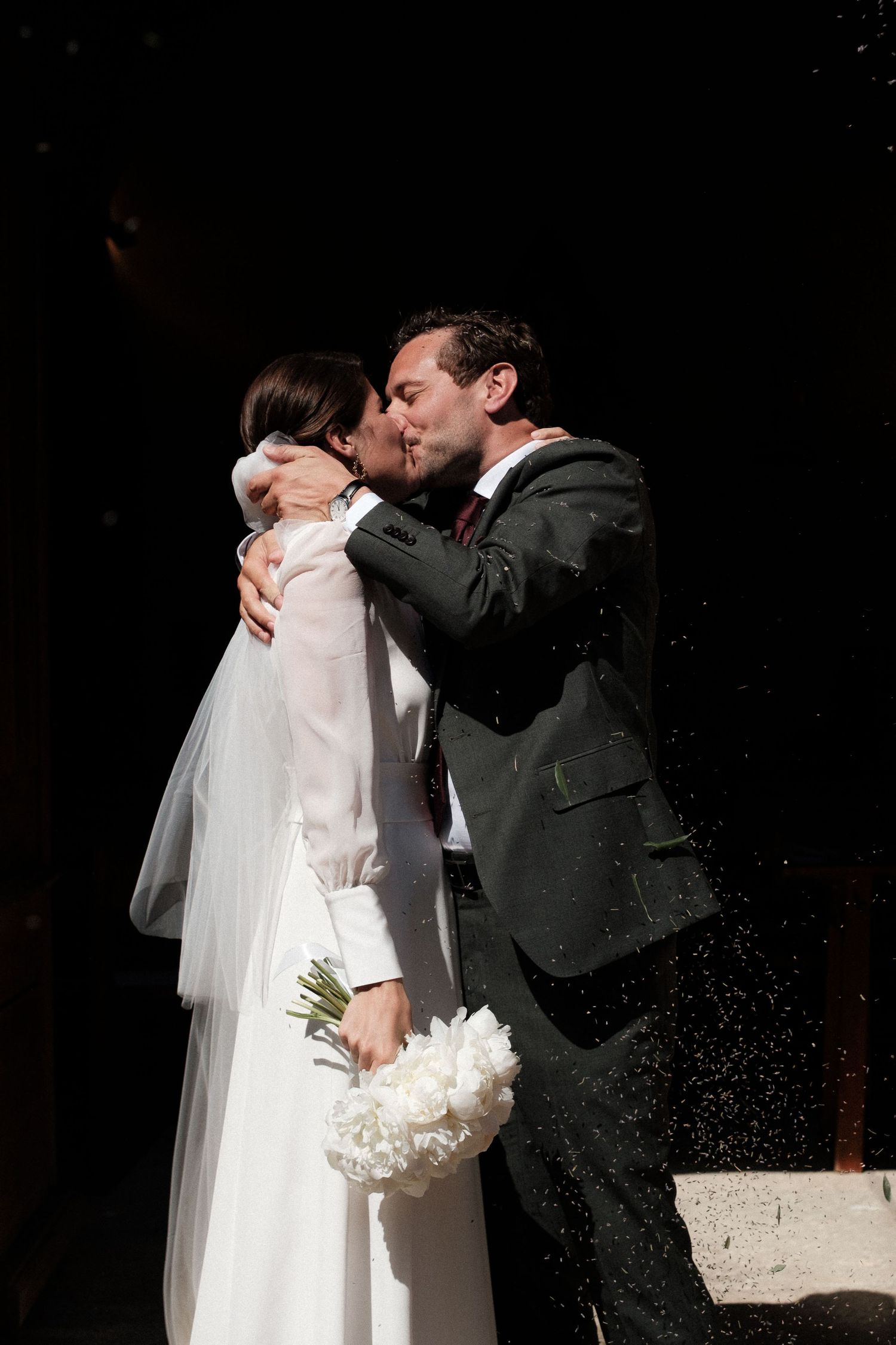 A bride and groom share a romantic kiss during their wedding ceremony against a dark background.