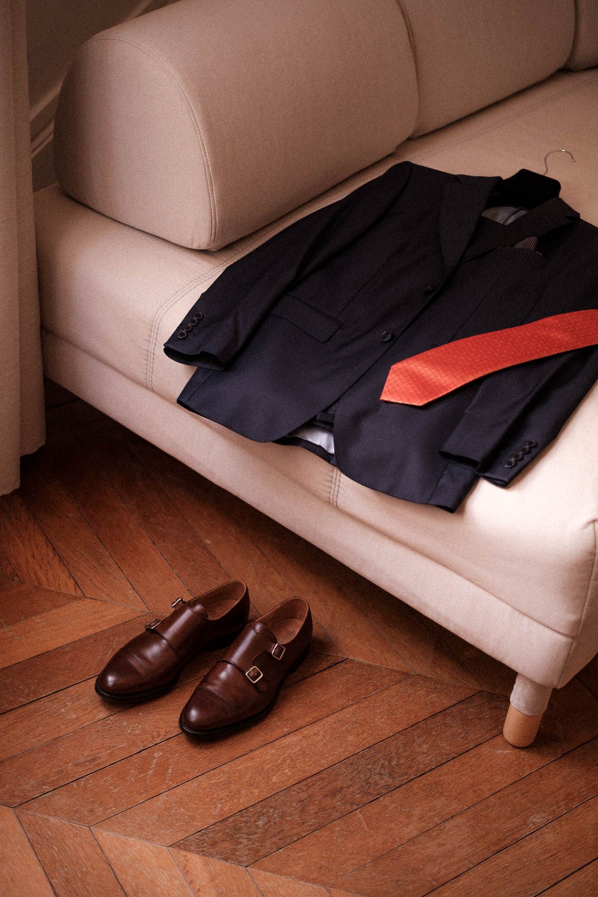 Navy suit jacket and brown leather shoes arranged beside a beige couch on wooden flooring.