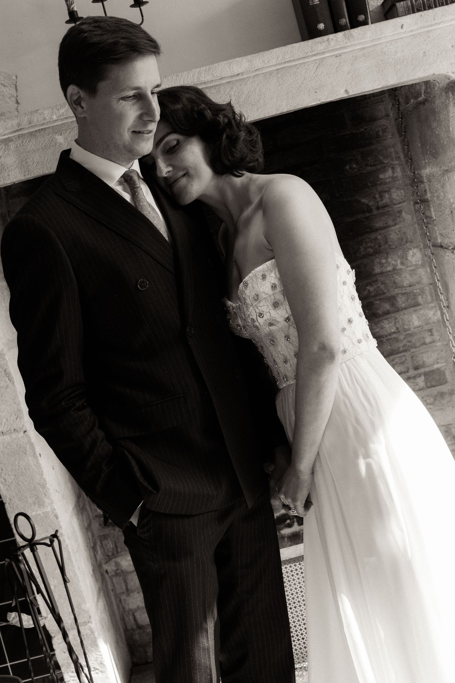 A black and white wedding photo of a couple in formal attire standing by a fireplace, sharing an intimate moment.