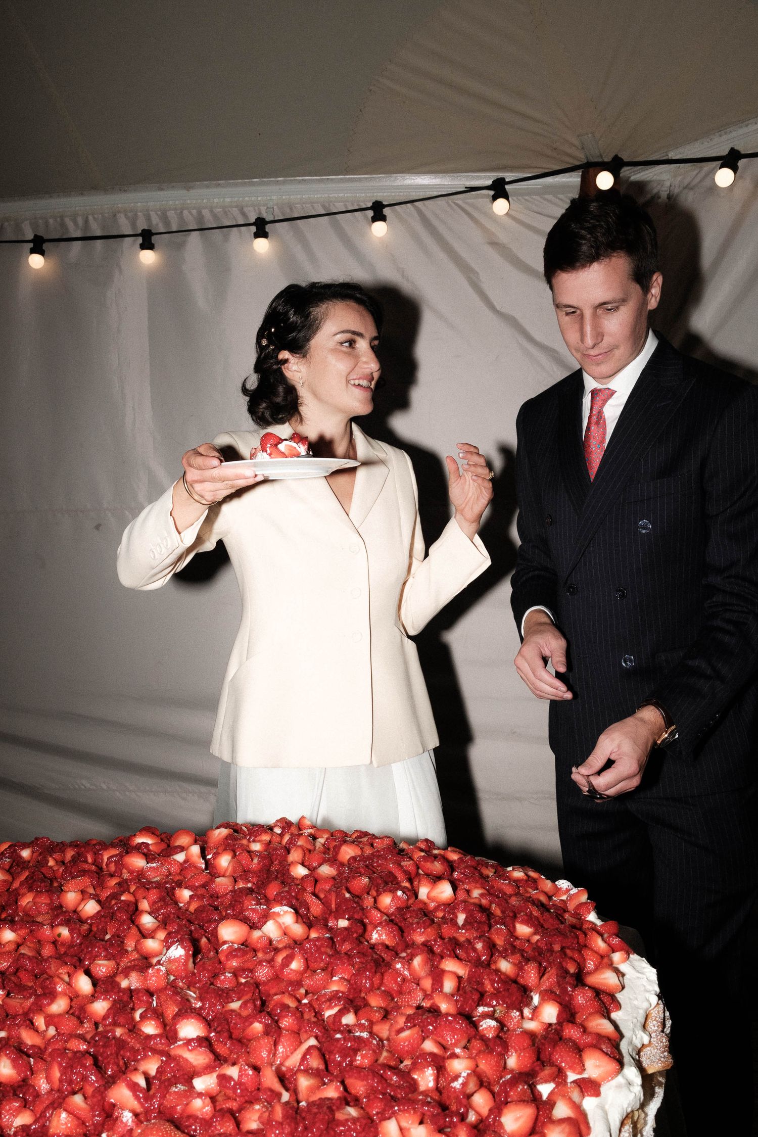 A couple cuts into a large red and white wedding cake decorated with fresh strawberries under string lights.