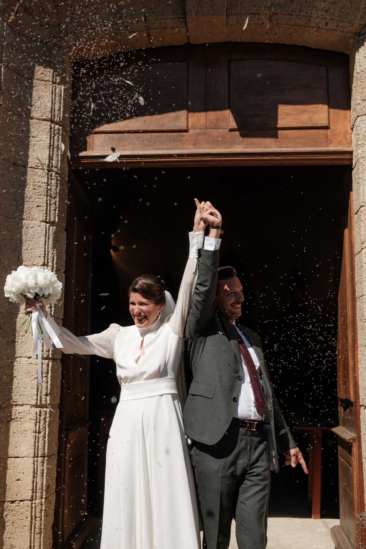 Newlyweds celebrate joyfully under stone archway with confetti raining down.