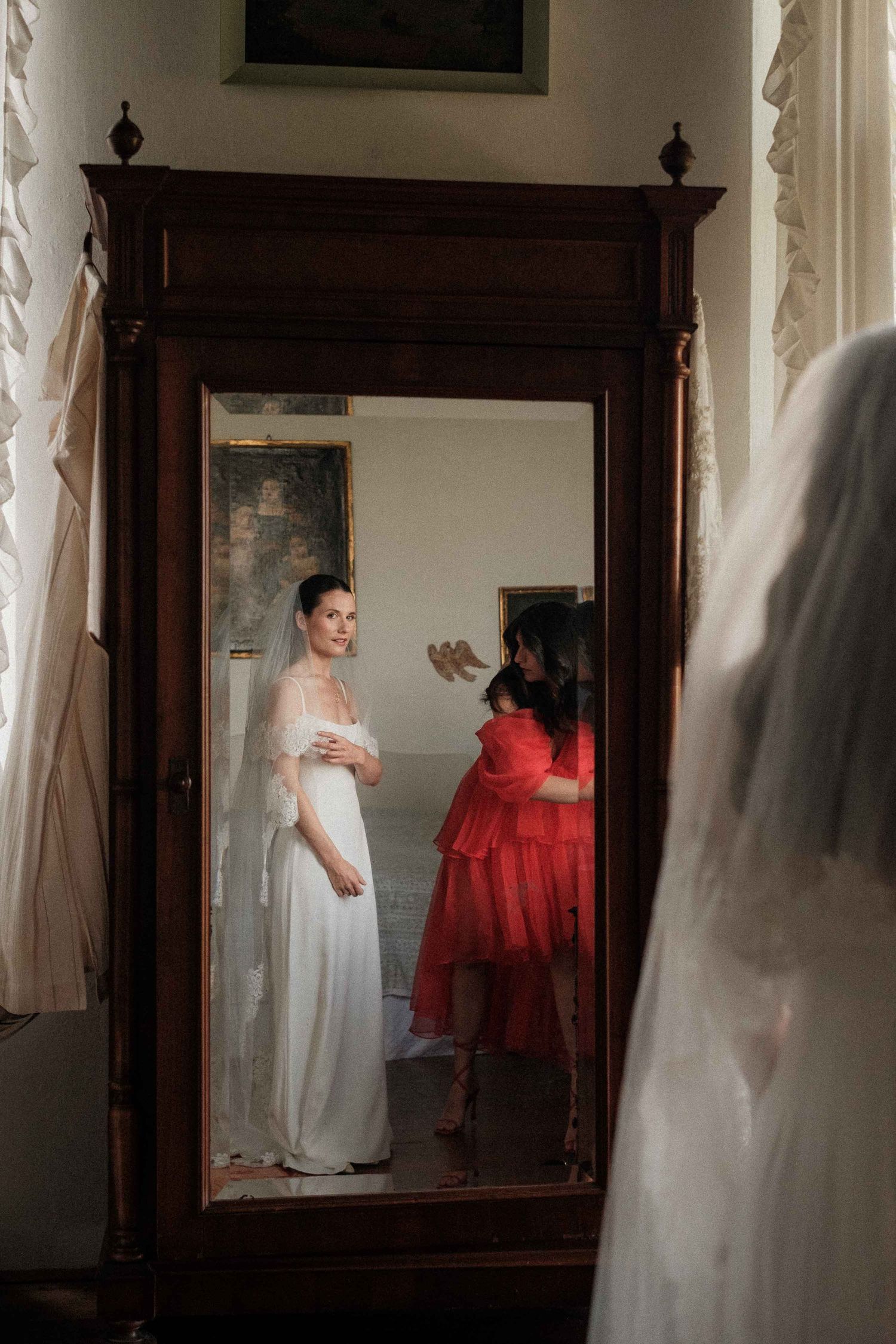 A figure in a white wedding dress stands before an ornate wooden mirror while someone in red adjusts the gown.