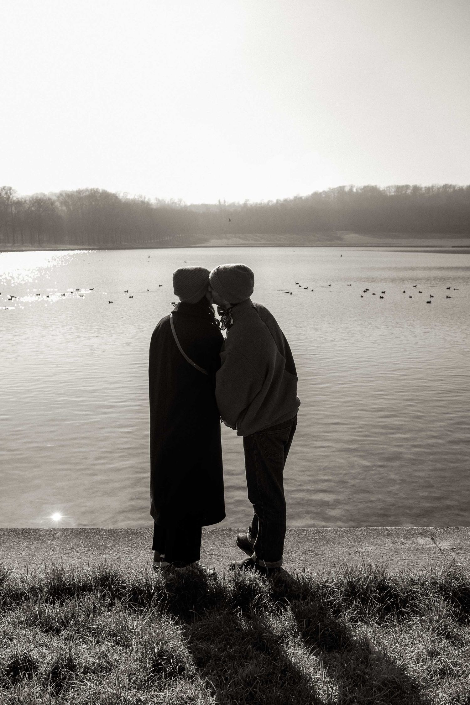 Two silhouetted figures stand close together by a misty lake with birds floating on the calm waters in black and white.