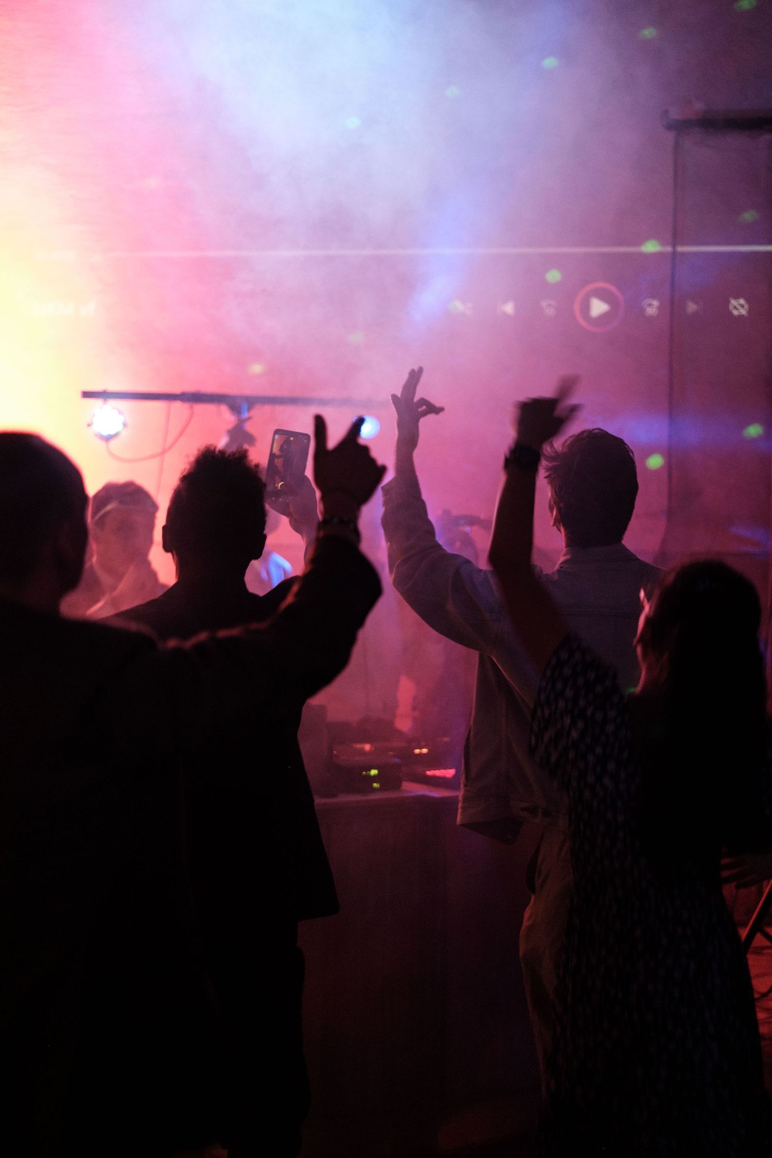 Silhouetted crowd with raised hands at a concert with colorful pink and purple stage lighting effects.
