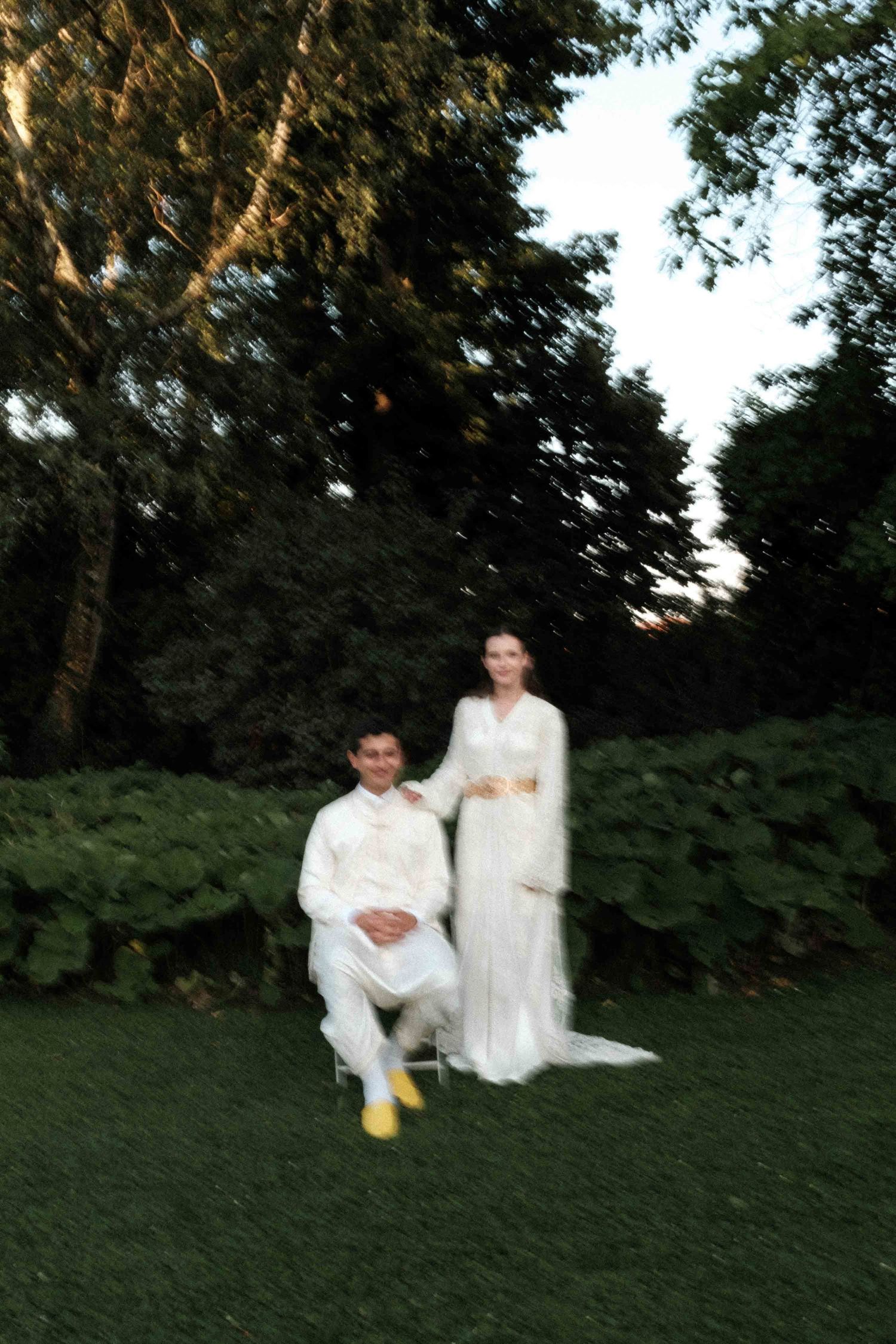 A couple in white outfits plays together on a grassy hill at dusk with trees in the background.