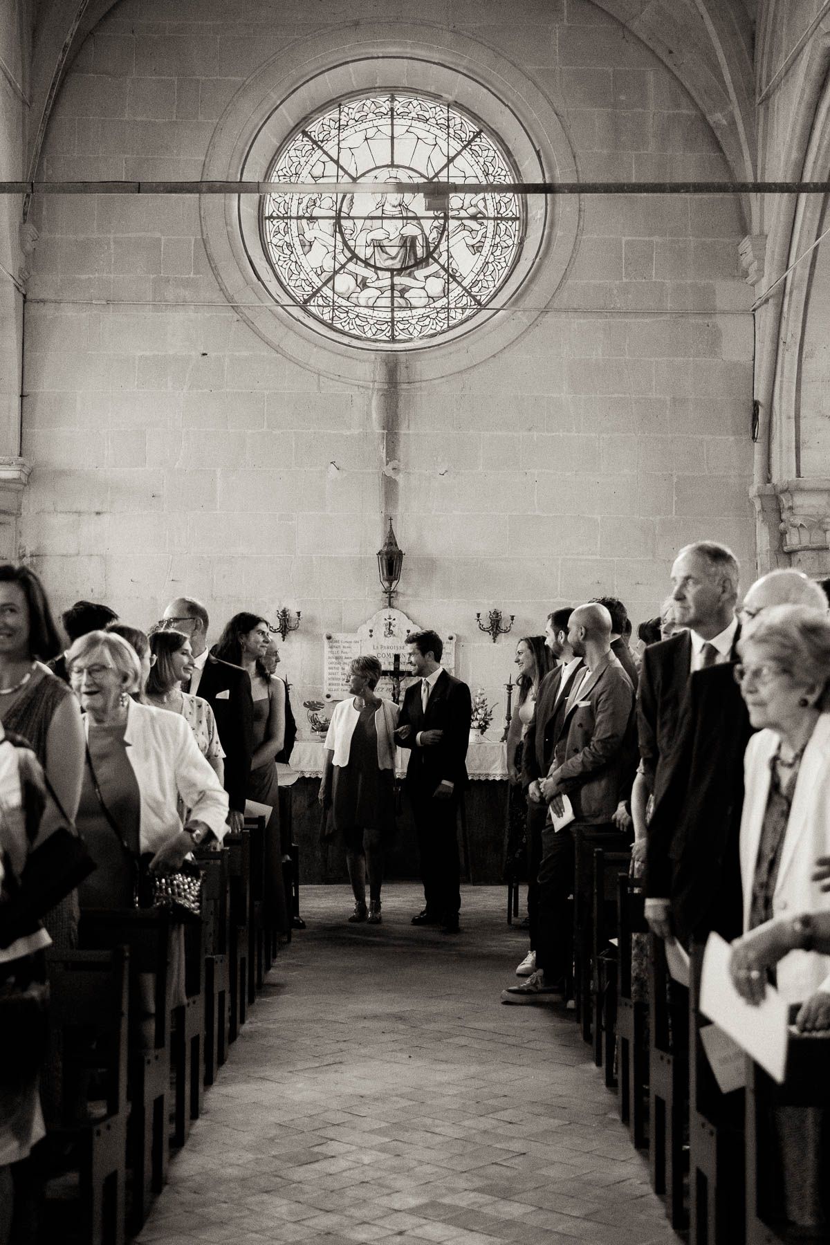 Wedding guests stand in church pews beneath a beautiful round stained glass window during a ceremony.