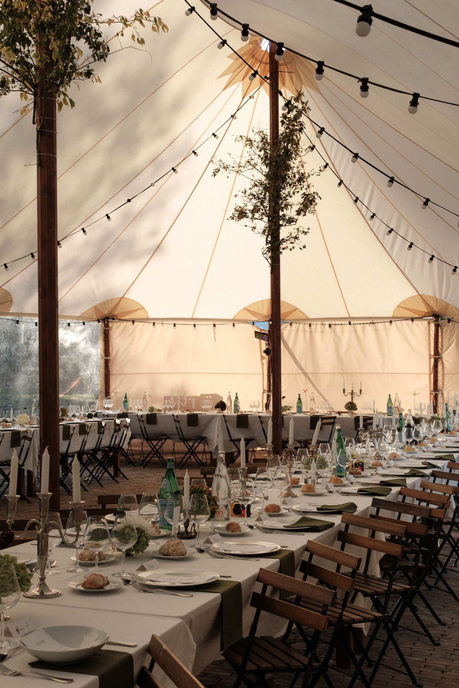 Wedding reception tent setup with string lights, wooden folding chairs and elegant table settings at sunset.