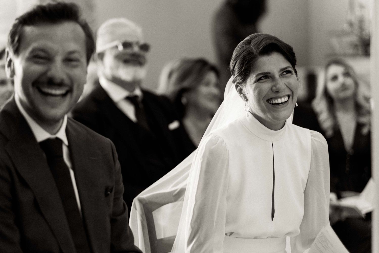 A joyful black and white wedding photograph capturing candid laughter between a bride in white and guests during the ceremony.