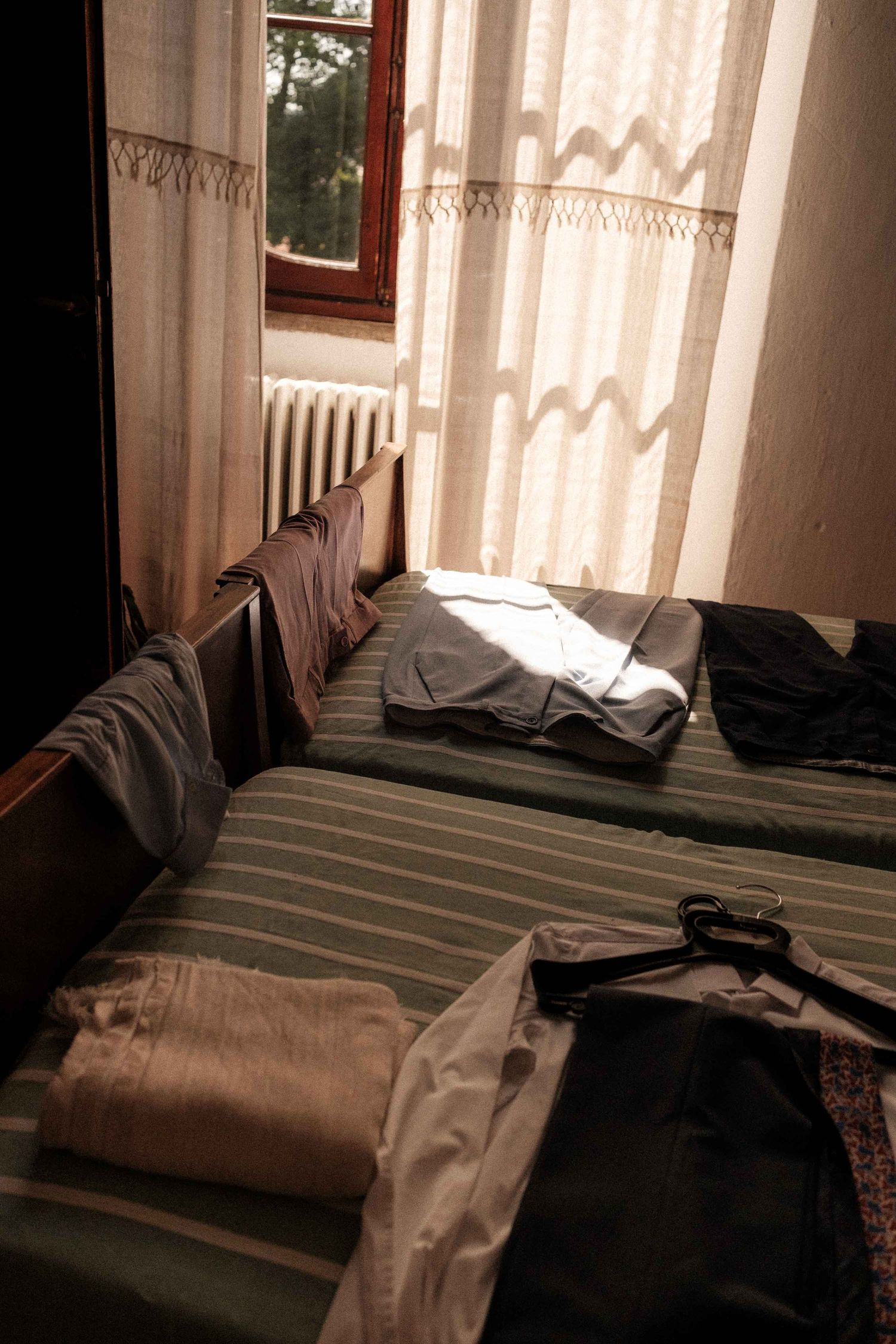Sunlight streams through white curtains onto an unmade bed with striped bedding in a cozy bedroom setting.