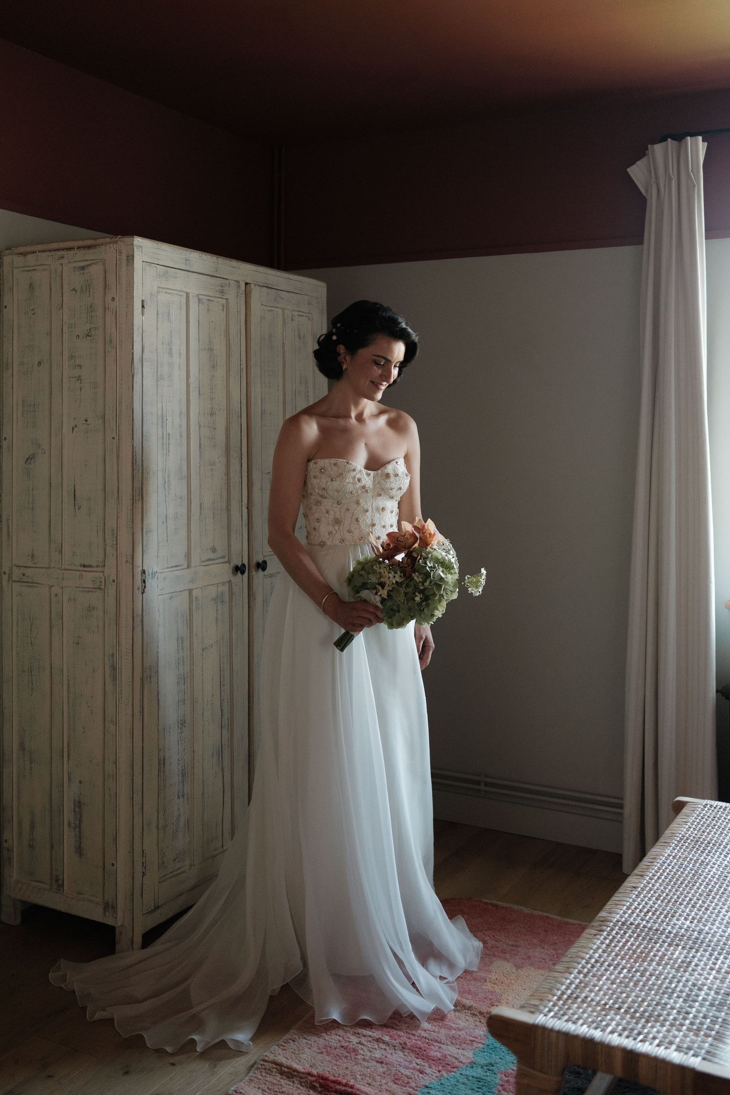 Elegant bridal portrait in flowing white gown against rustic wooden wardrobe and soft window light.