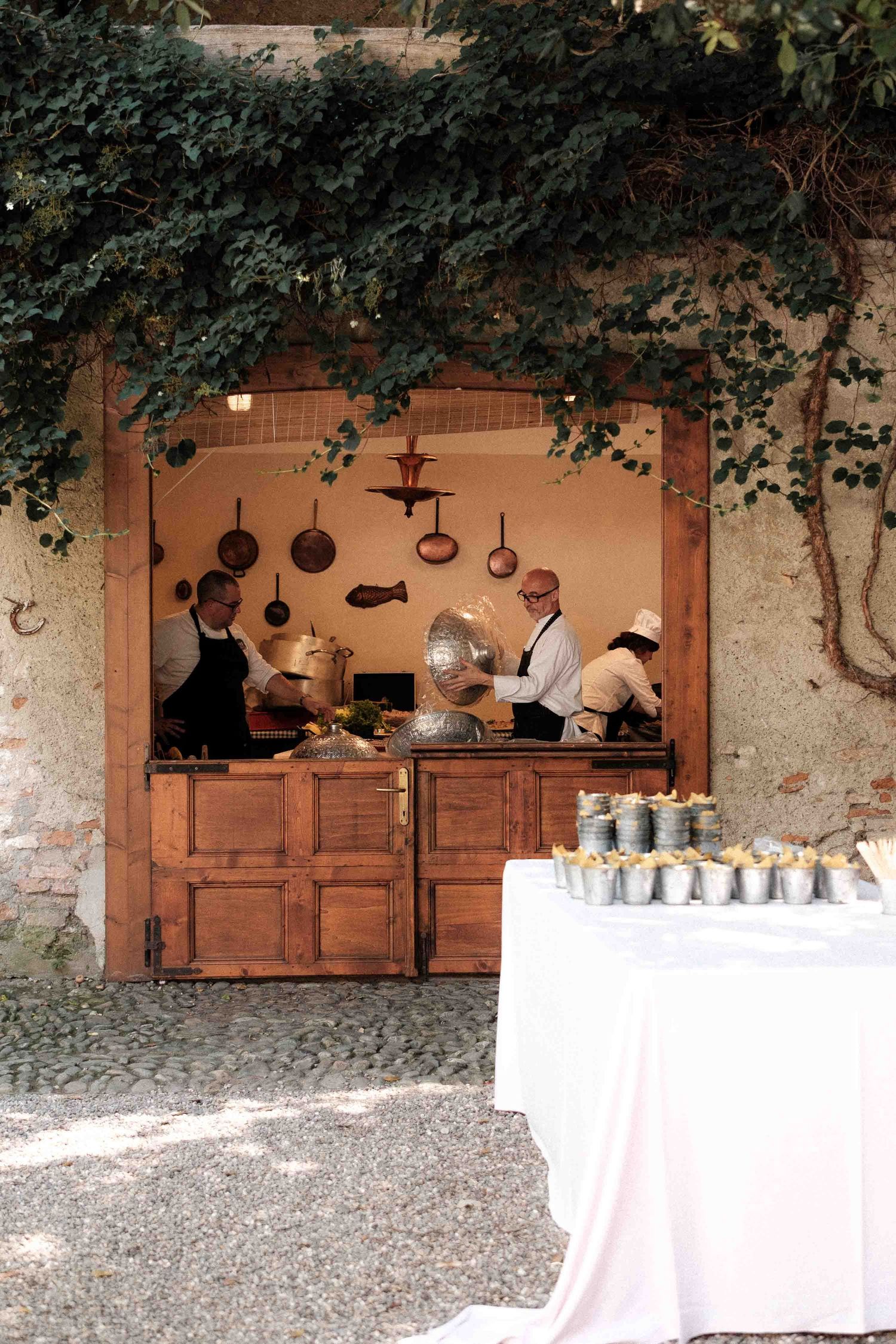 Rustic outdoor kitchen setup with copper pots hanging and white-clothed tables arranged against stone walls.