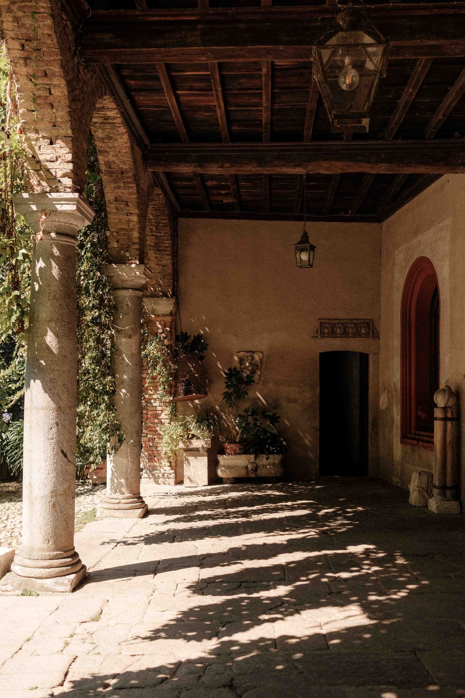 Sunlight casts dramatic shadows through stone columns along a rustic covered walkway in an old Mediterranean building.