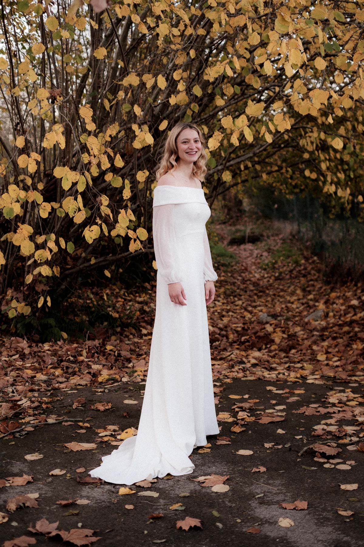 A person in a flowing white wedding dress poses against a backdrop of golden autumn leaves and fallen foliage.