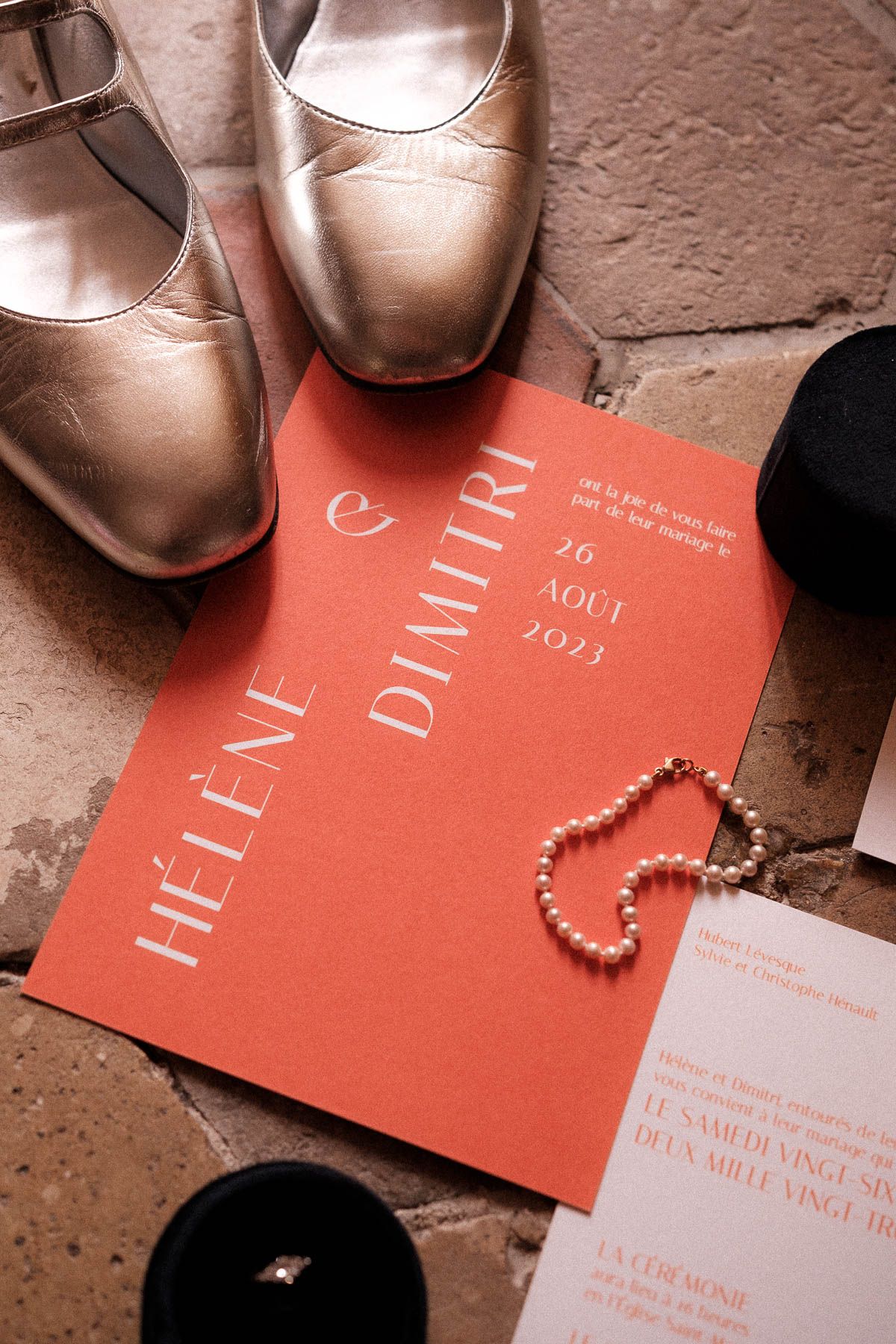 Red wedding invitations with gold lettering displayed alongside shiny metallic shoes and pearl beaded jewelry.