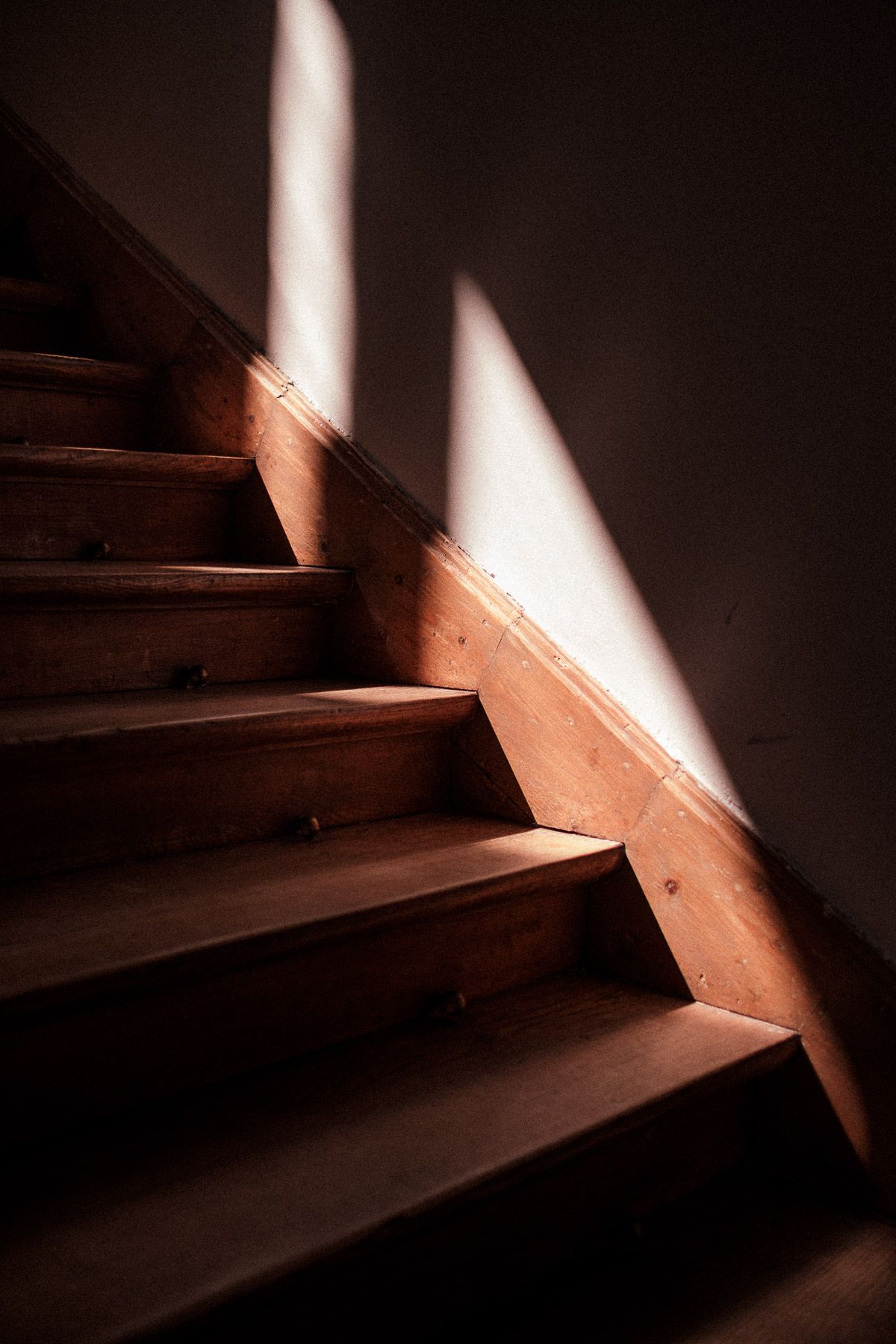 Dramatic sunlight beams illuminate wooden stairs in a dark stairwell creating striking angular shadows.