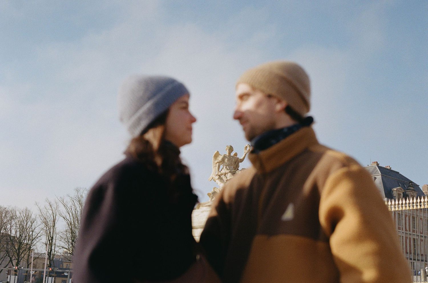 A couple in winter coats walks together in a romantic outdoor setting with bare trees in the background.