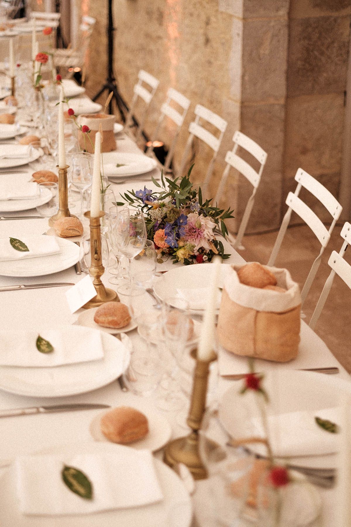 Elegant table setting with white plates, small bread bowls, and floral centerpieces in a rustic stone venue.