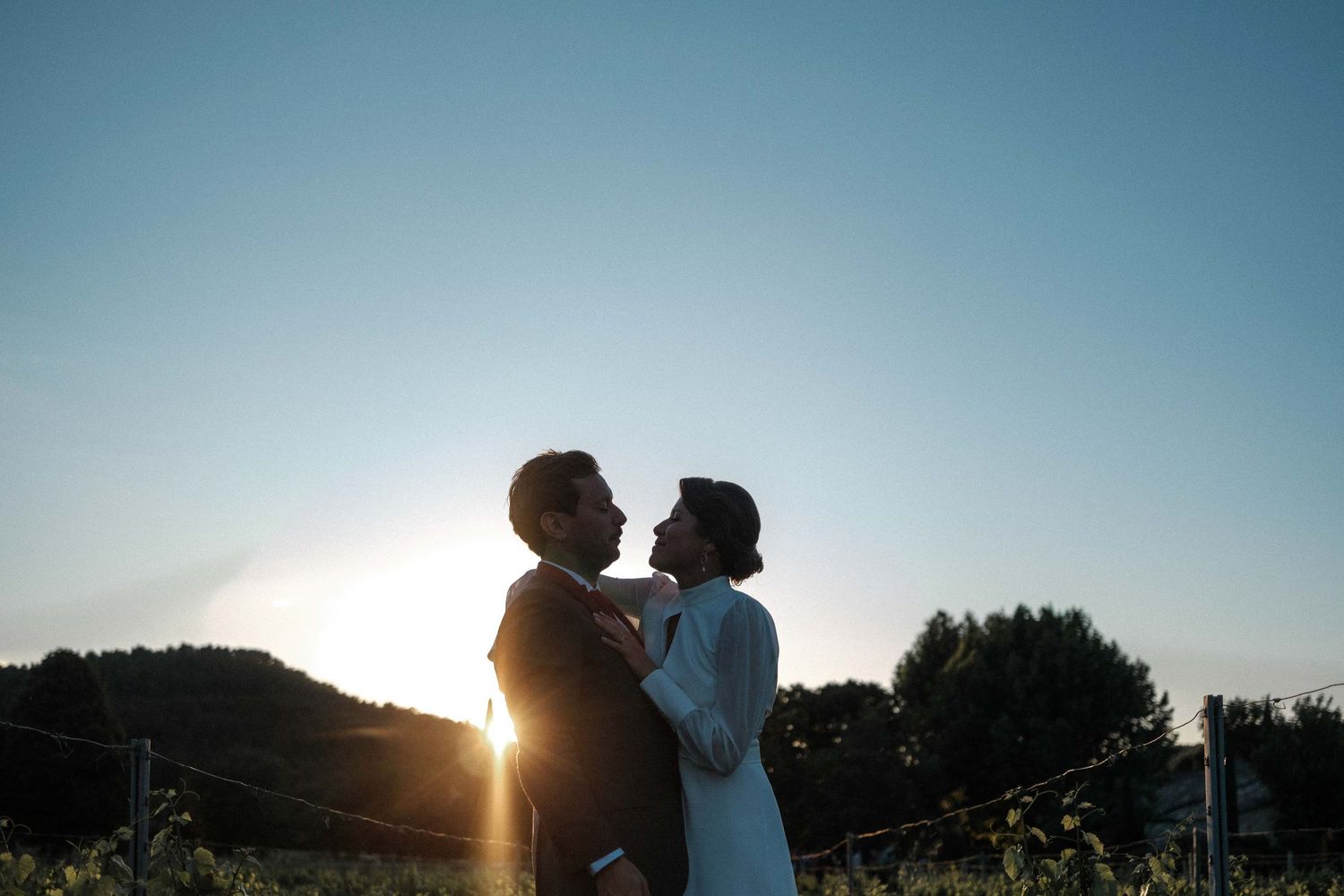 A silhouetted couple embraces in front of a sunset with trees in the background.