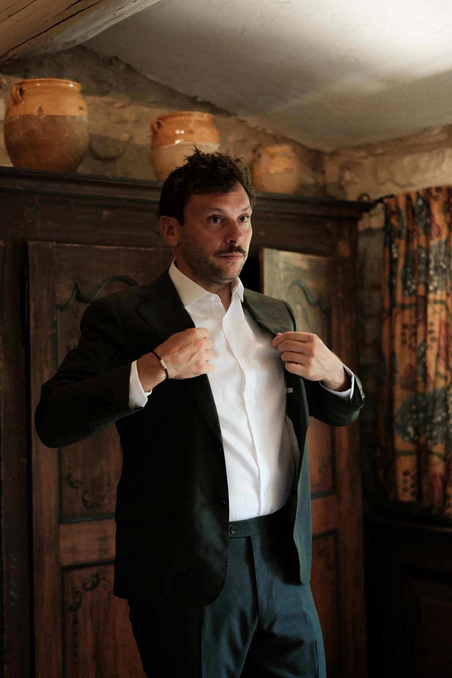 A person in a dark suit and white shirt adjusts their collar in a dimly lit room with floral wallpaper.