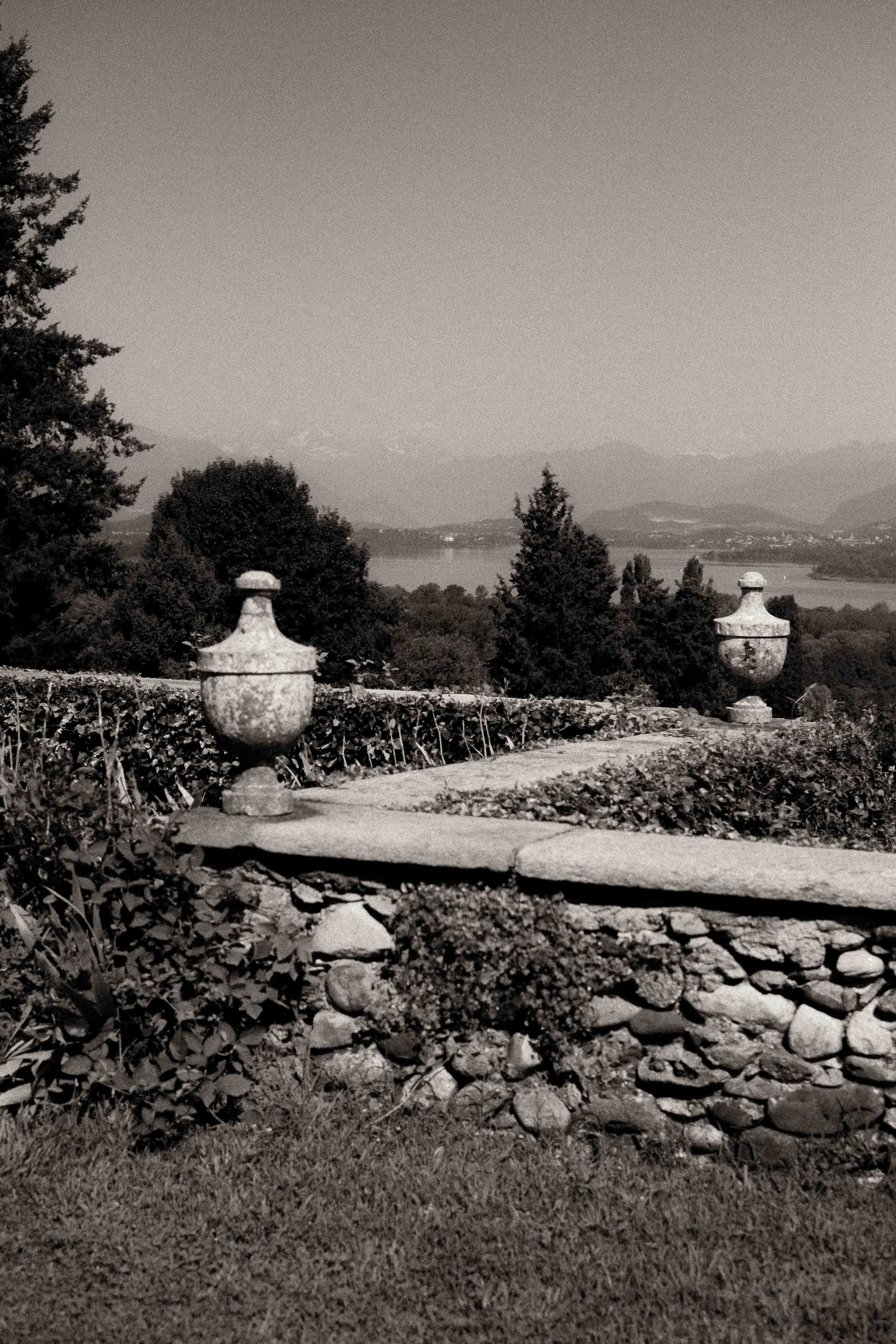 Black and white landscape view of decorative stone urns atop a rustic garden wall overlooking a distant lake and mountains.
