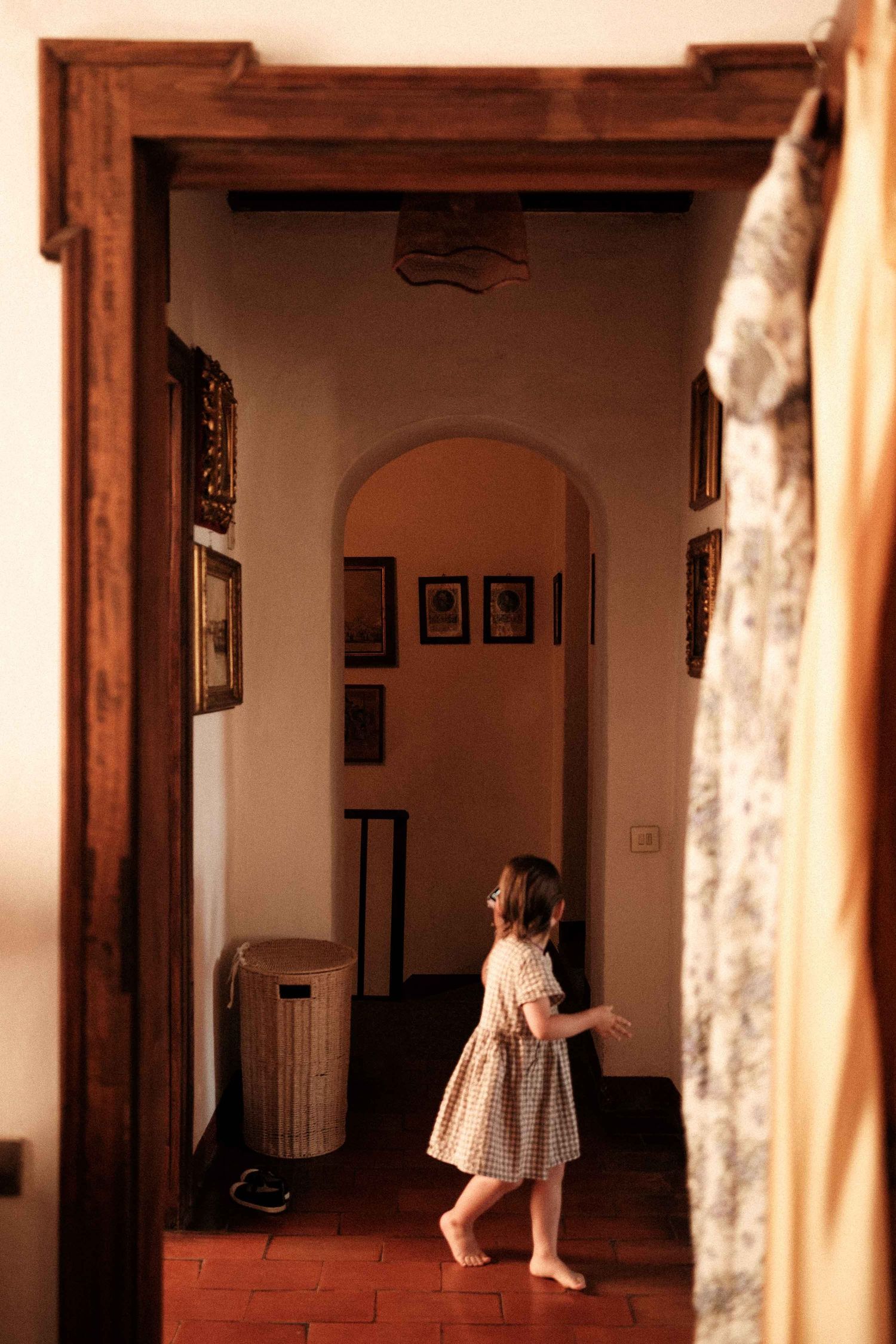A small figure in a dress stands in a dimly lit hallway with terracotta floors and framed artwork on the walls.