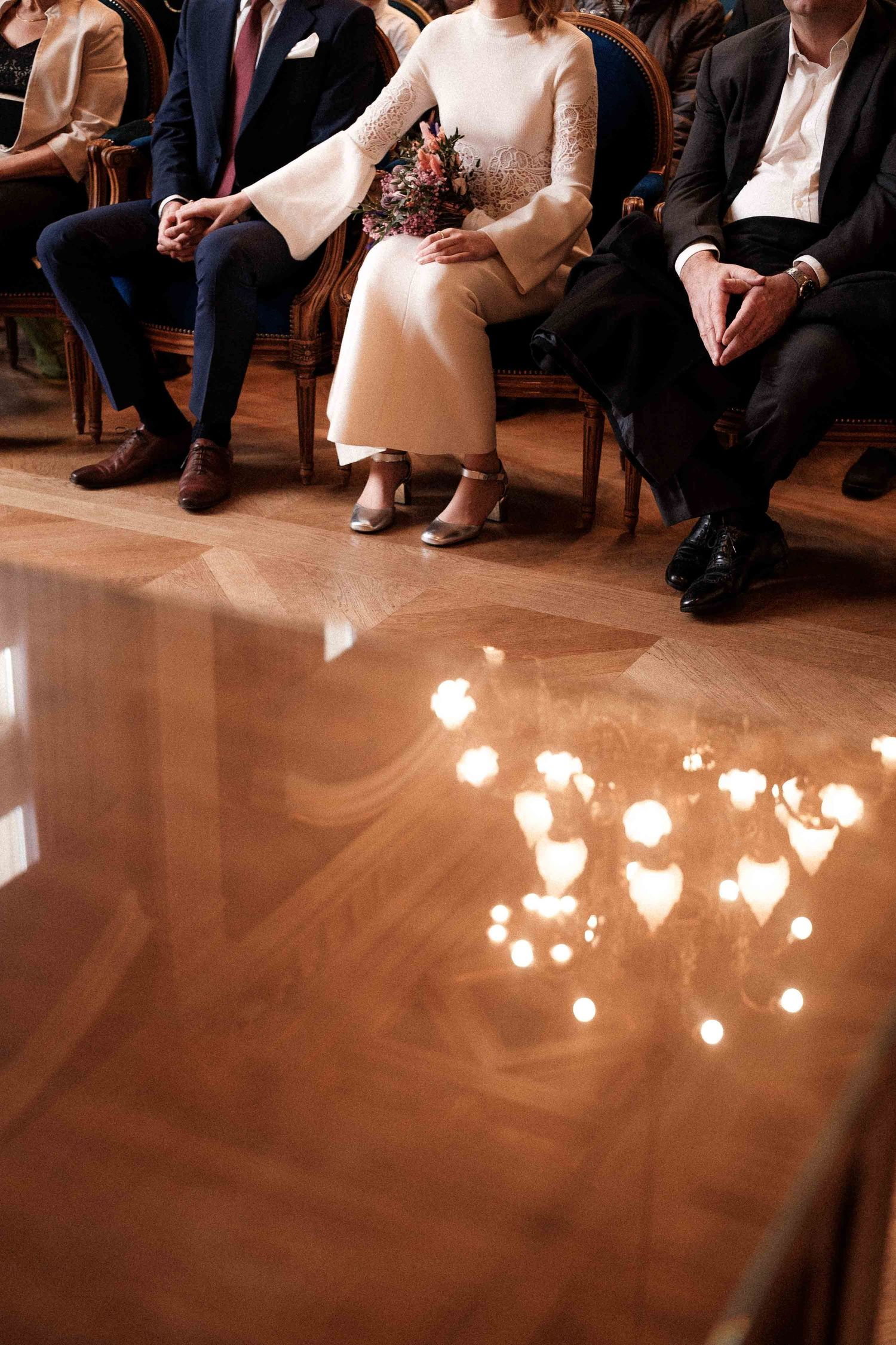 Soft candlelight reflects on polished wooden floor at formal event with seated guests in elegant attire.