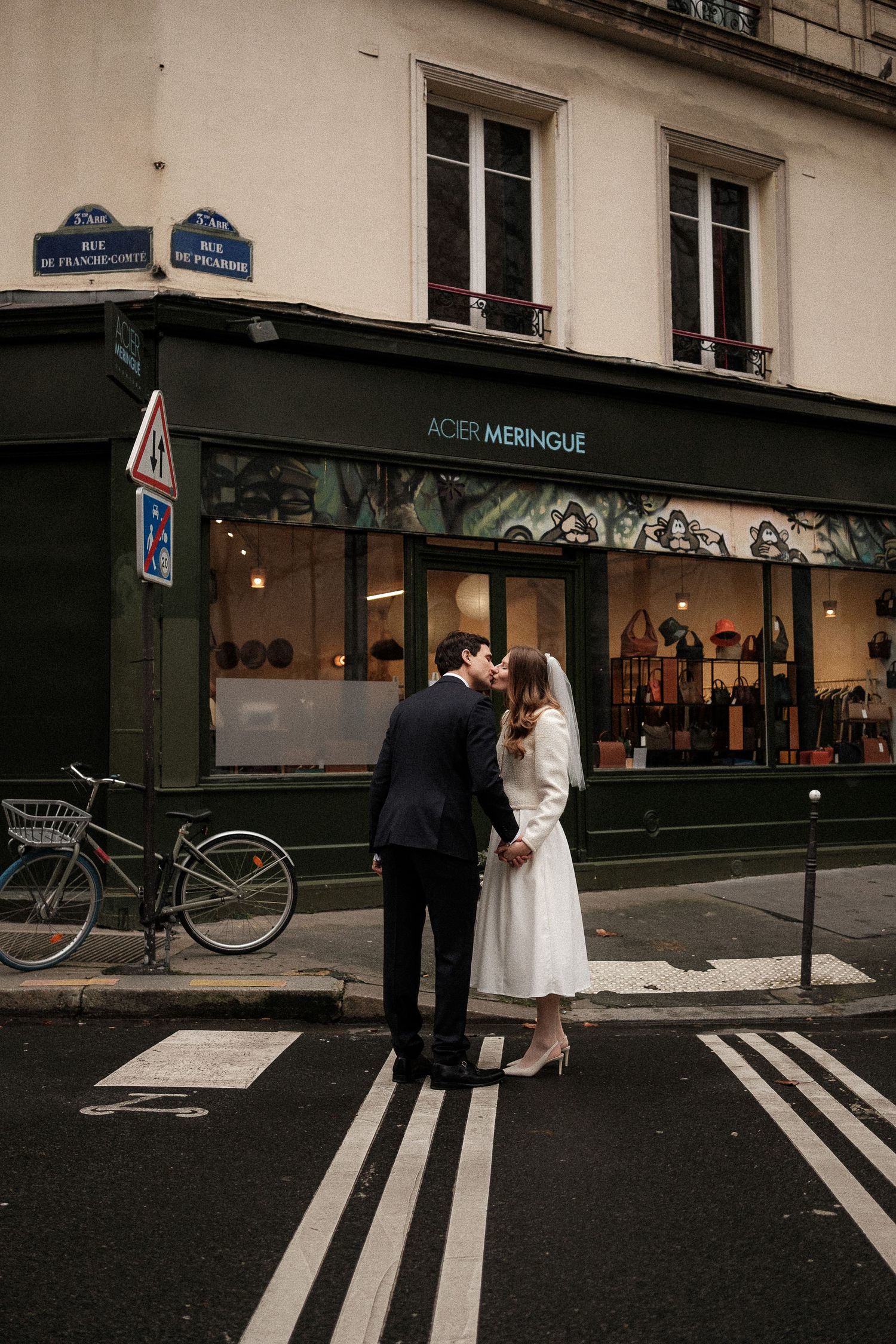 A couple in formal attire stands at a crosswalk in front of a Parisian storefront with a bicycle visible nearby.