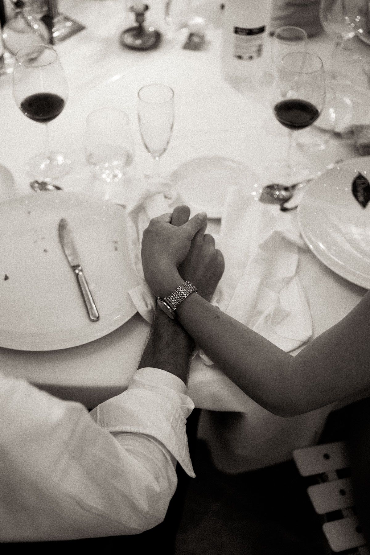 A black and white image of two hands clasped together on a formal dining table with wine glasses and place settings.
