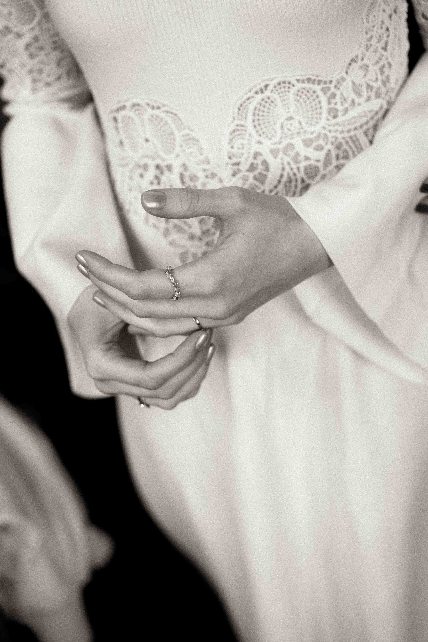 Close-up black and white photo of hands with wedding rings against delicate white lace dress detail.
