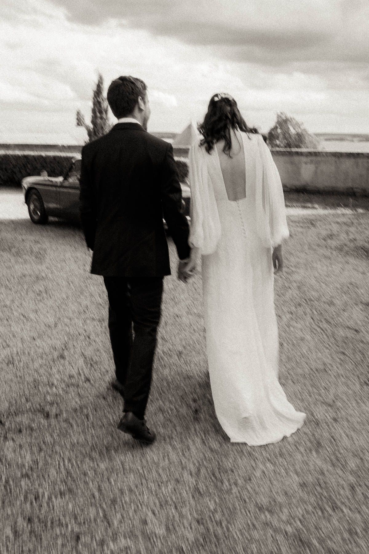 A bride and groom walk together on grass in black and white, with her flowing dress and veil captured from behind.