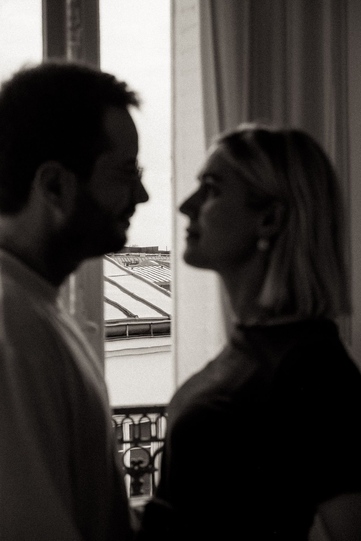 A romantic black and white silhouette of a couple facing each other by a window with rooftops visible outside.