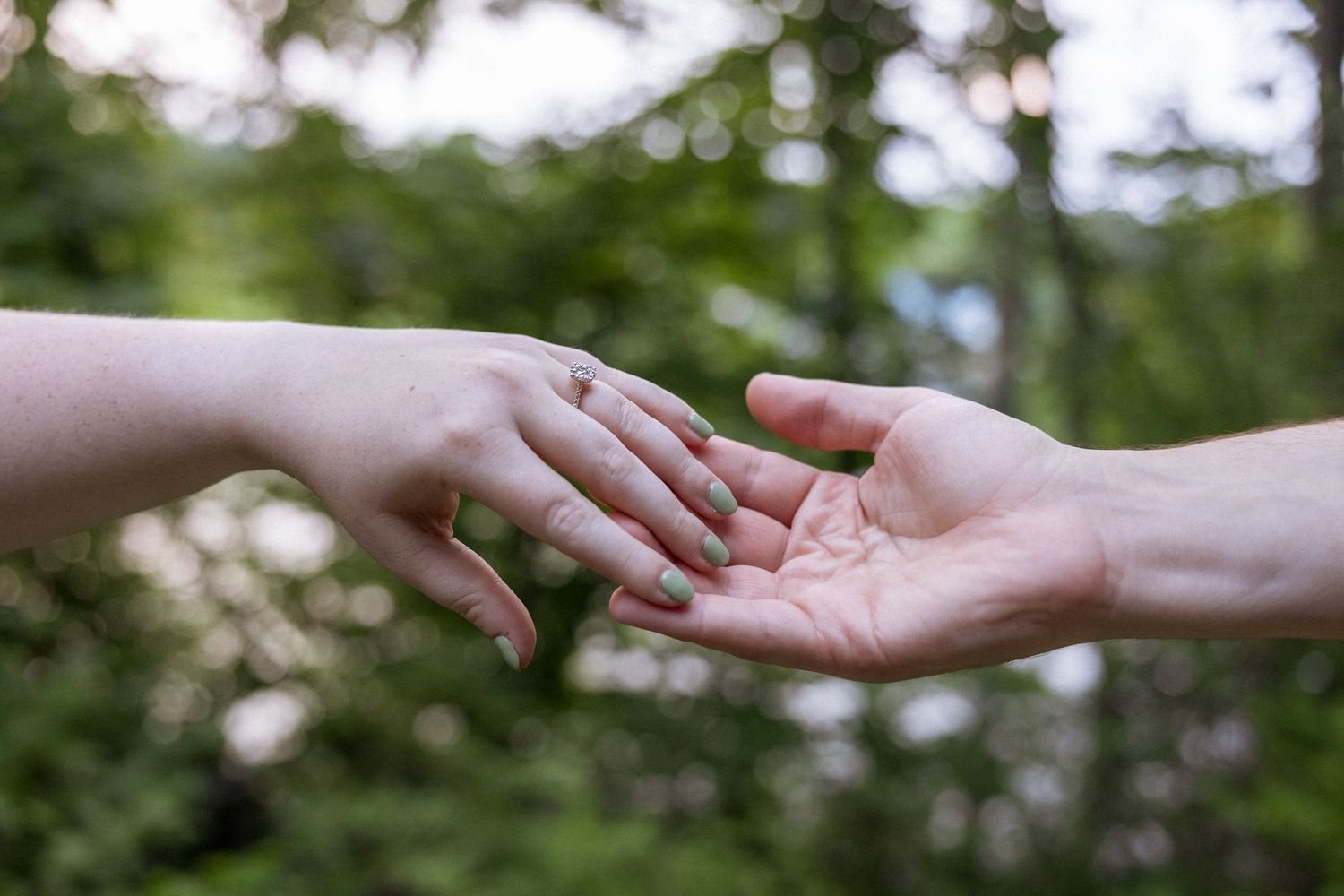 Two hands gently touching outdoors, one wearing a diamond engagement ring, with a blurred green forest background.