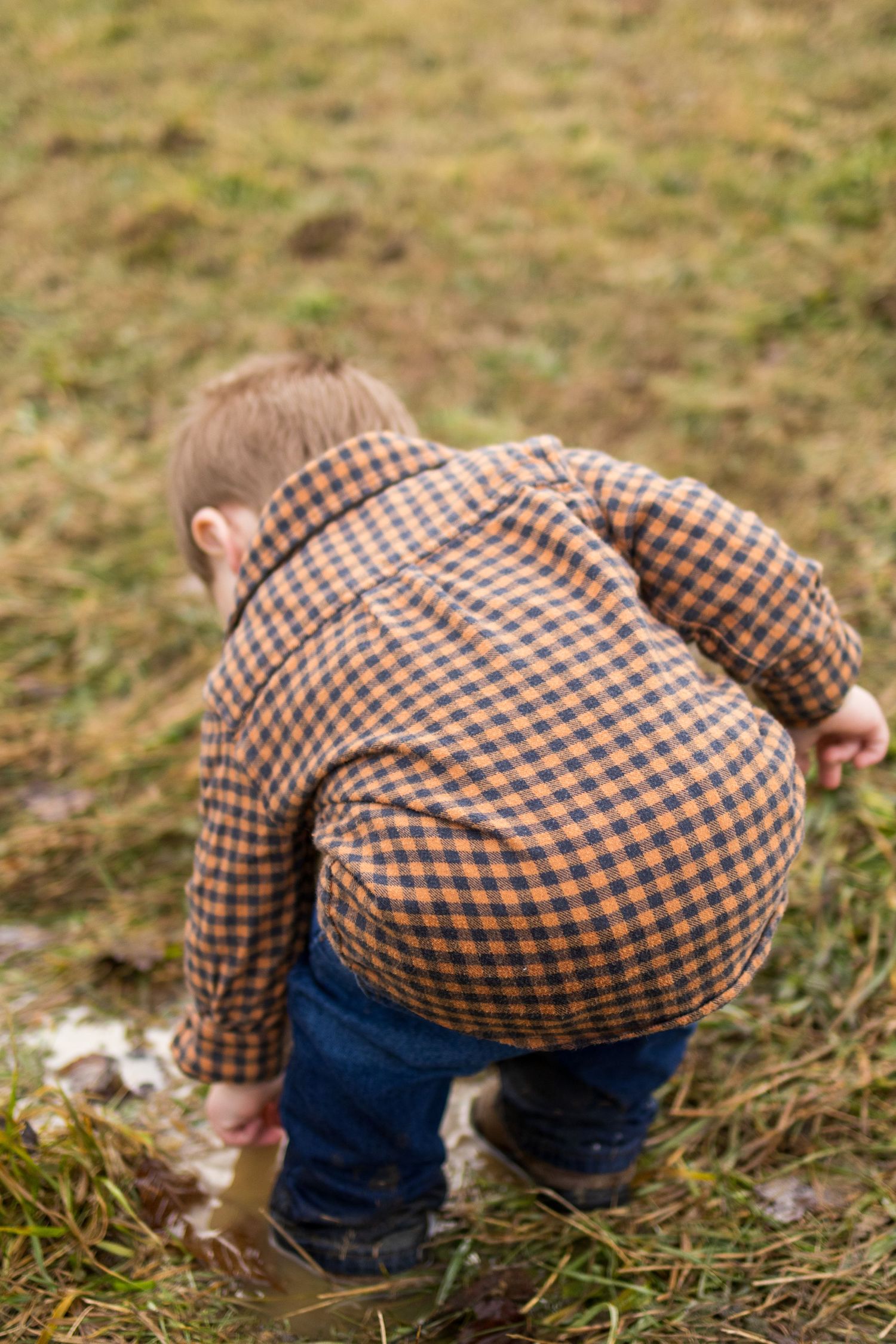 A young boy in a plaid shirt and jeans bends down to explore the ground outdoors on a grassy field.