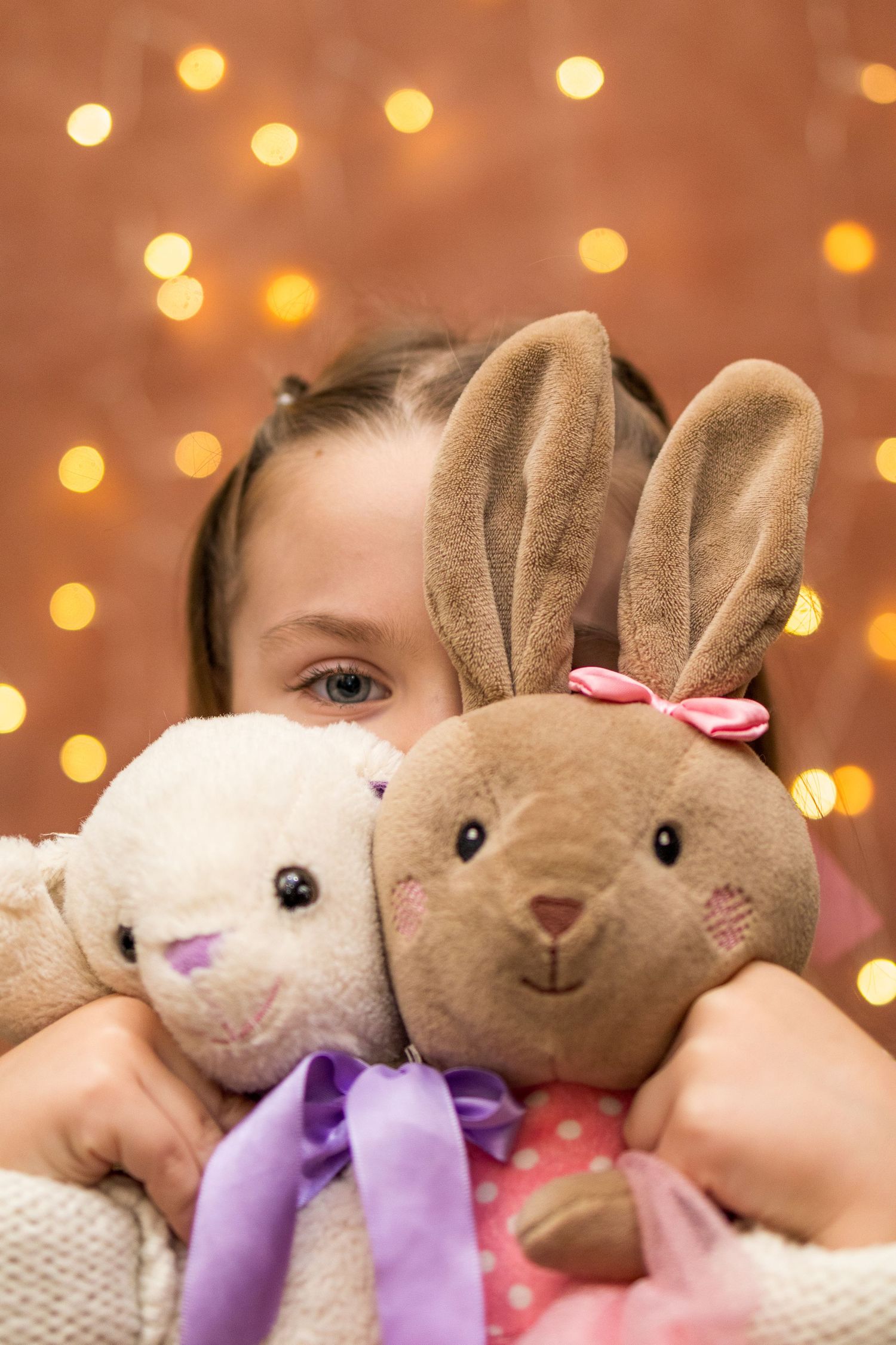 Young girl peeks over two stuffed bunny rabbits with bokeh lights in background.