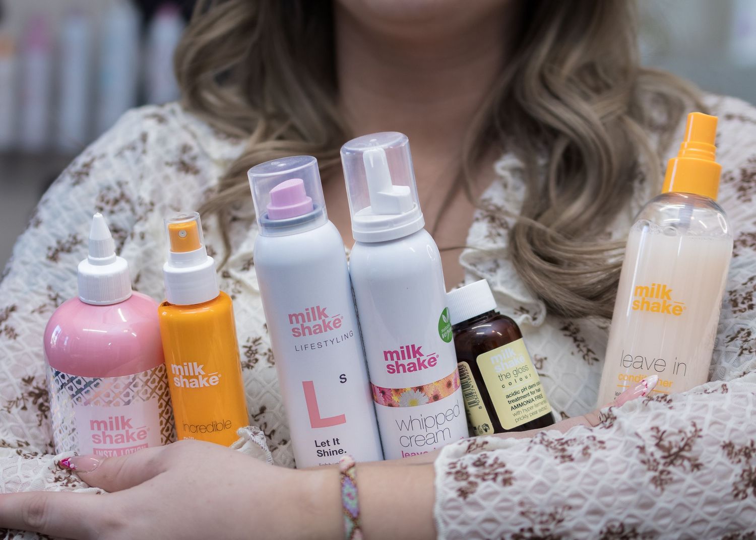Woman holding multiple Milk Shake hair care products including sprays and mousses arranged in a row.