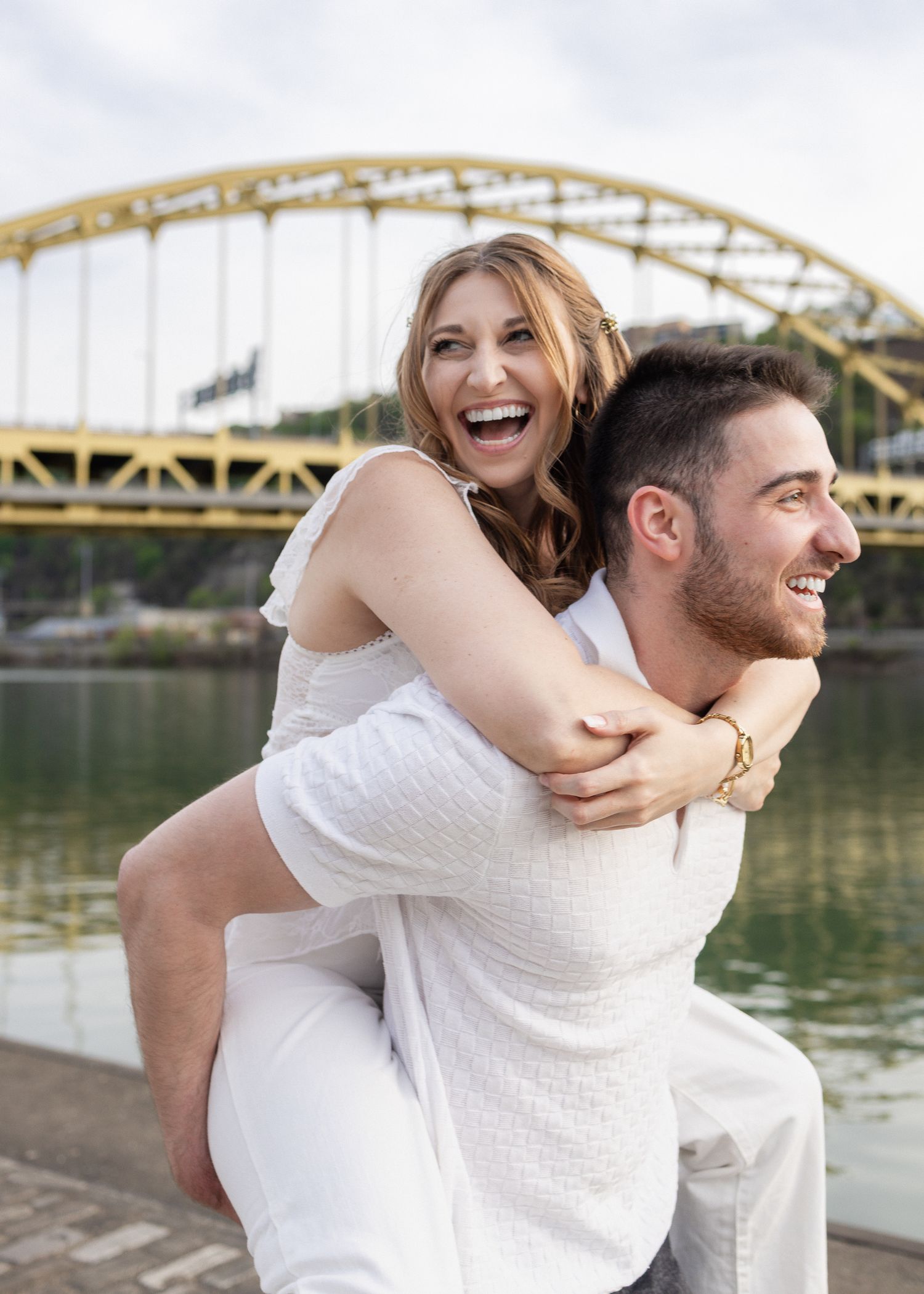 Happy couple in white outfits sharing a piggyback ride by a river with a yellow bridge backdrop.