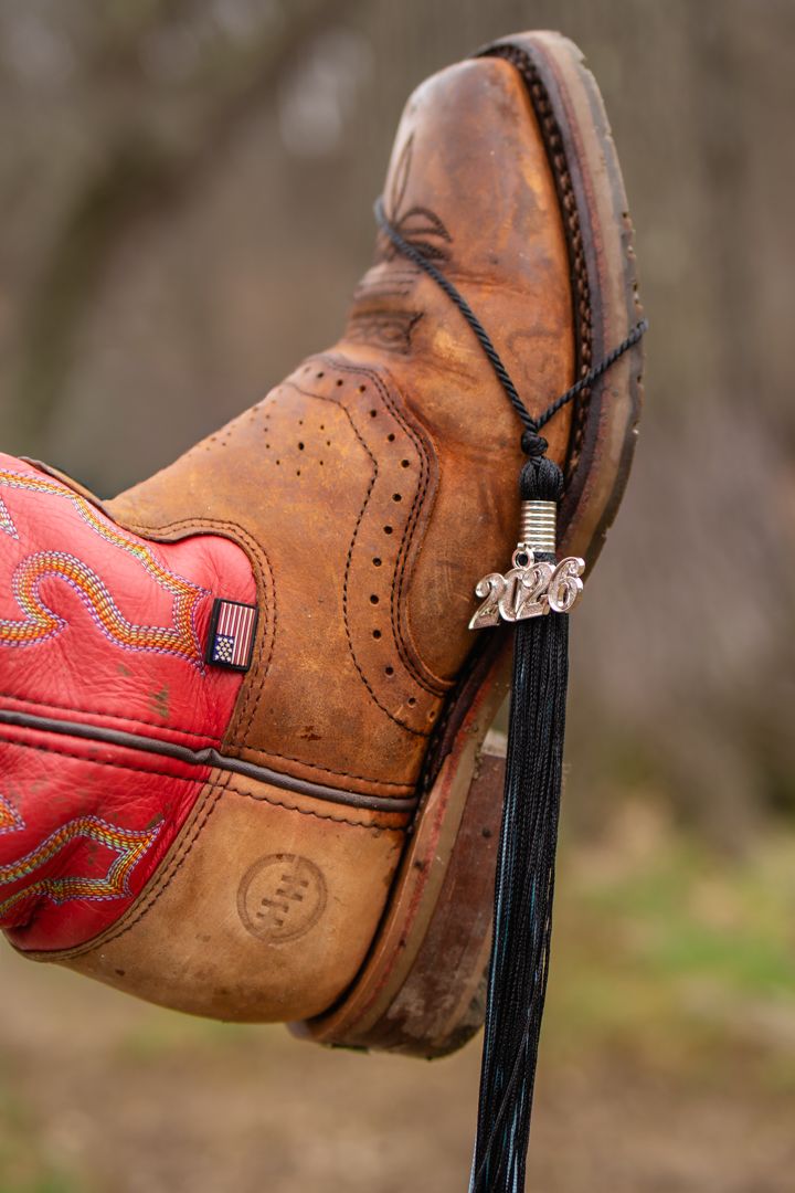 Brown cowboy boot with red stitching, American flag patch, and a 2026 graduation tassel charm, propped up outdoors.