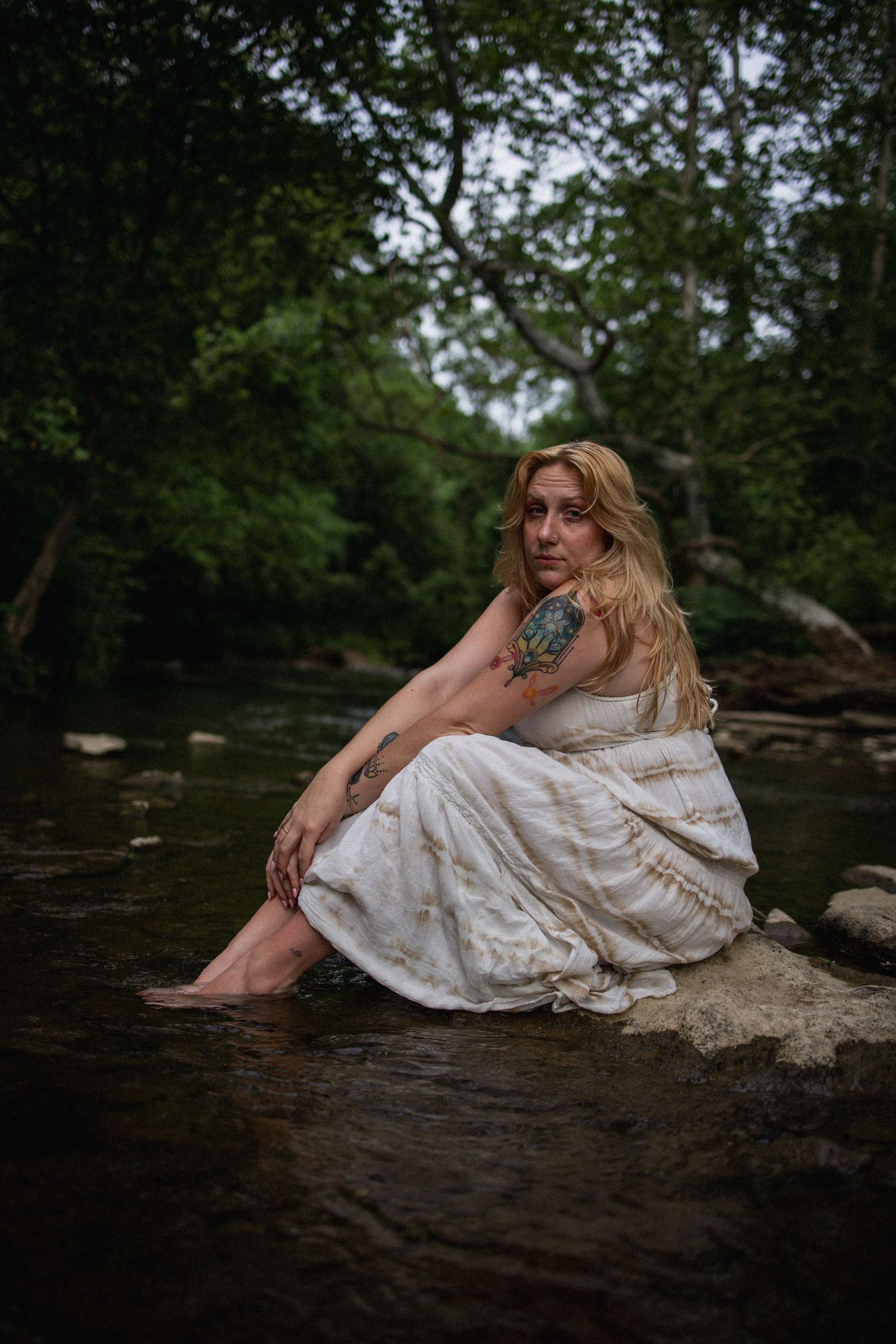 Blonde woman with tattoos in white dress sitting on rocks by a forest stream.