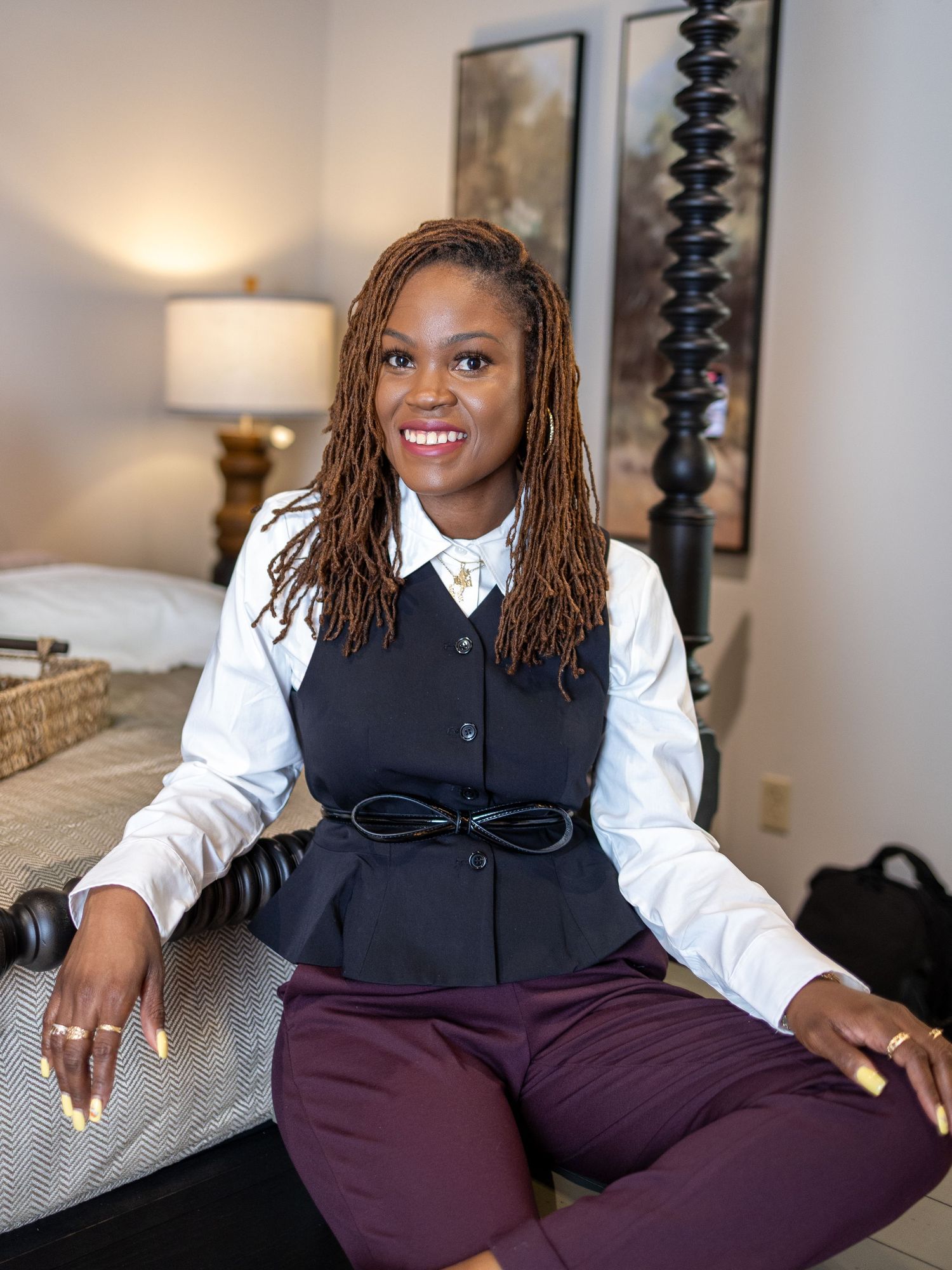 Professional woman in navy vest and white blouse sitting on bed, smiling confidently in a well-decorated room.