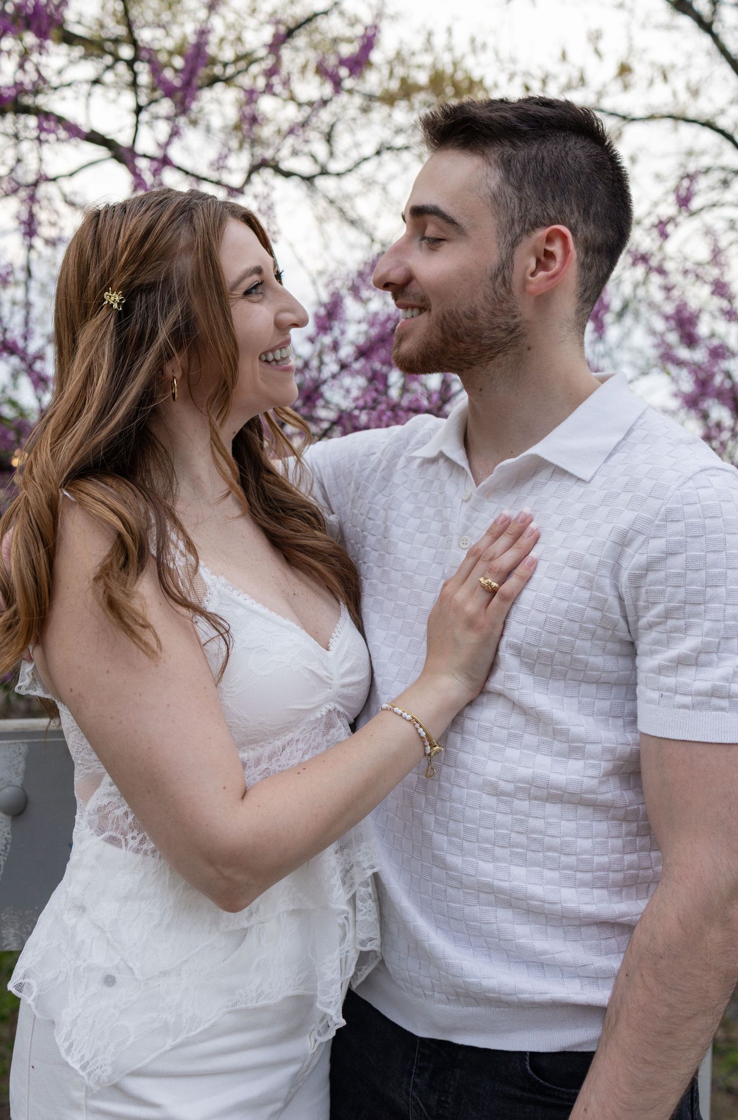 Couple smiling at each other outdoors with blooming pink trees, woman in white dress with engagement ring.
